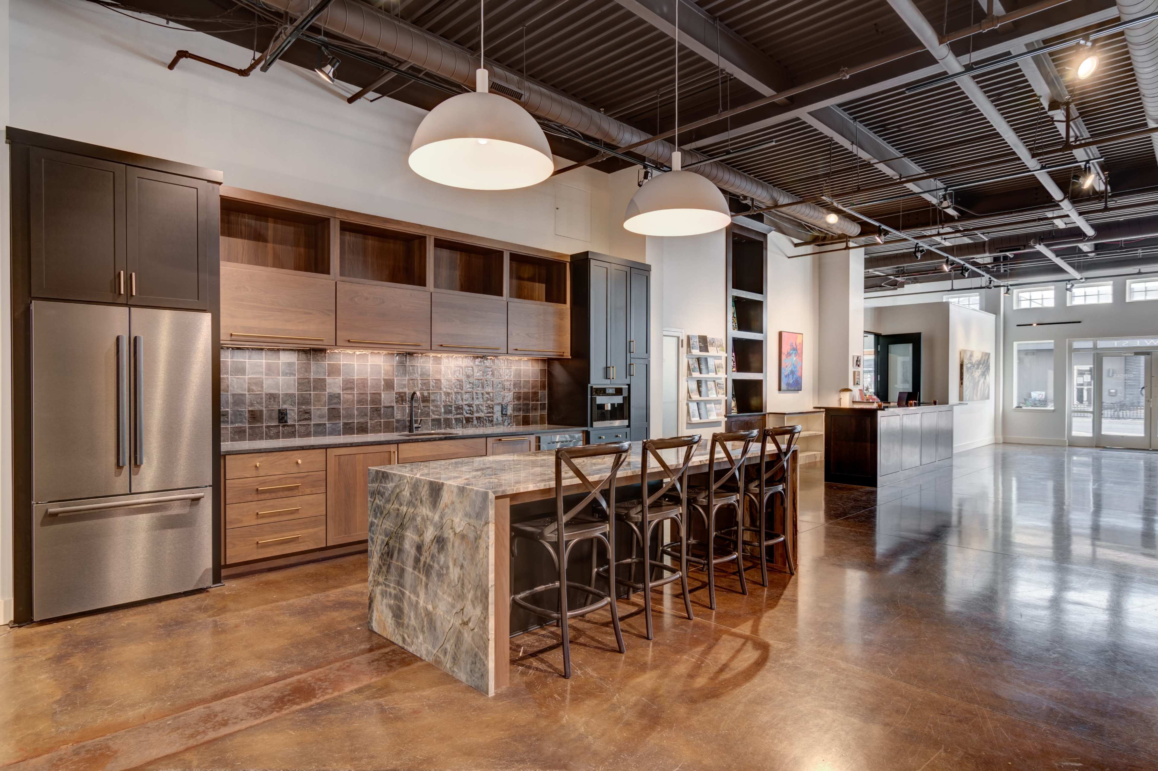 A modern kitchen with a large island and dark cabinetry, featuring a sleek stainless steel refrigerator and polished concrete flooring.