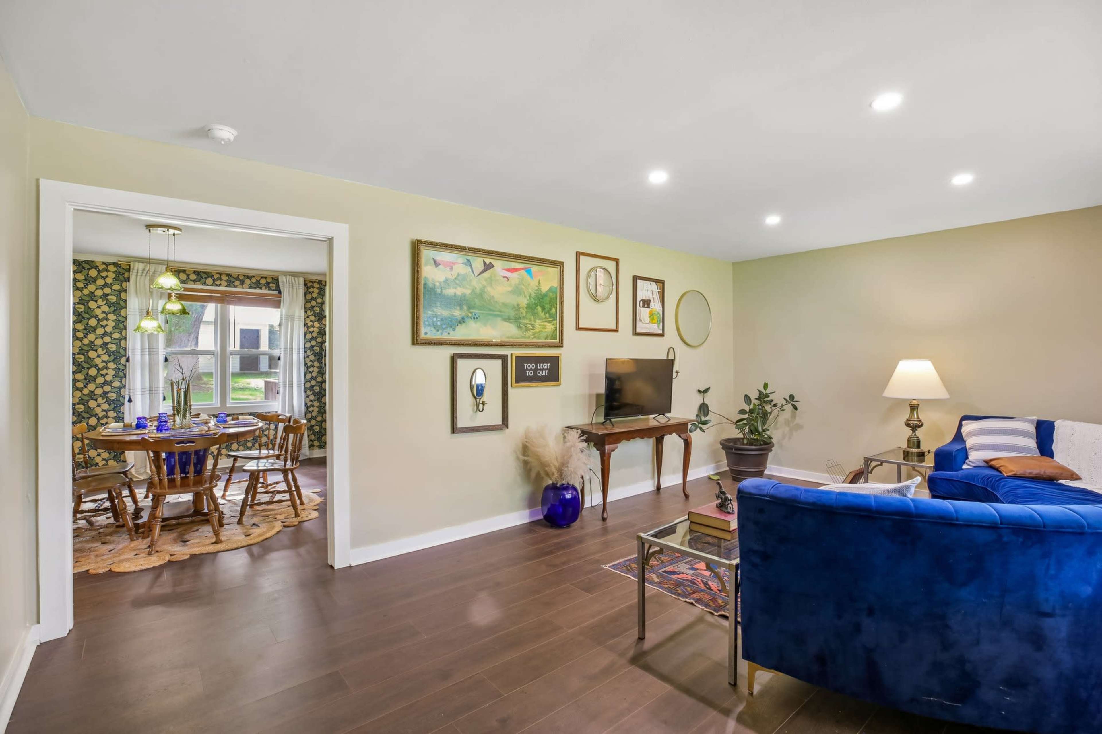 The image shows a living room with a blue couch, a television, and framed artwork, connecting to a dining area featuring a round wooden table and chairs.