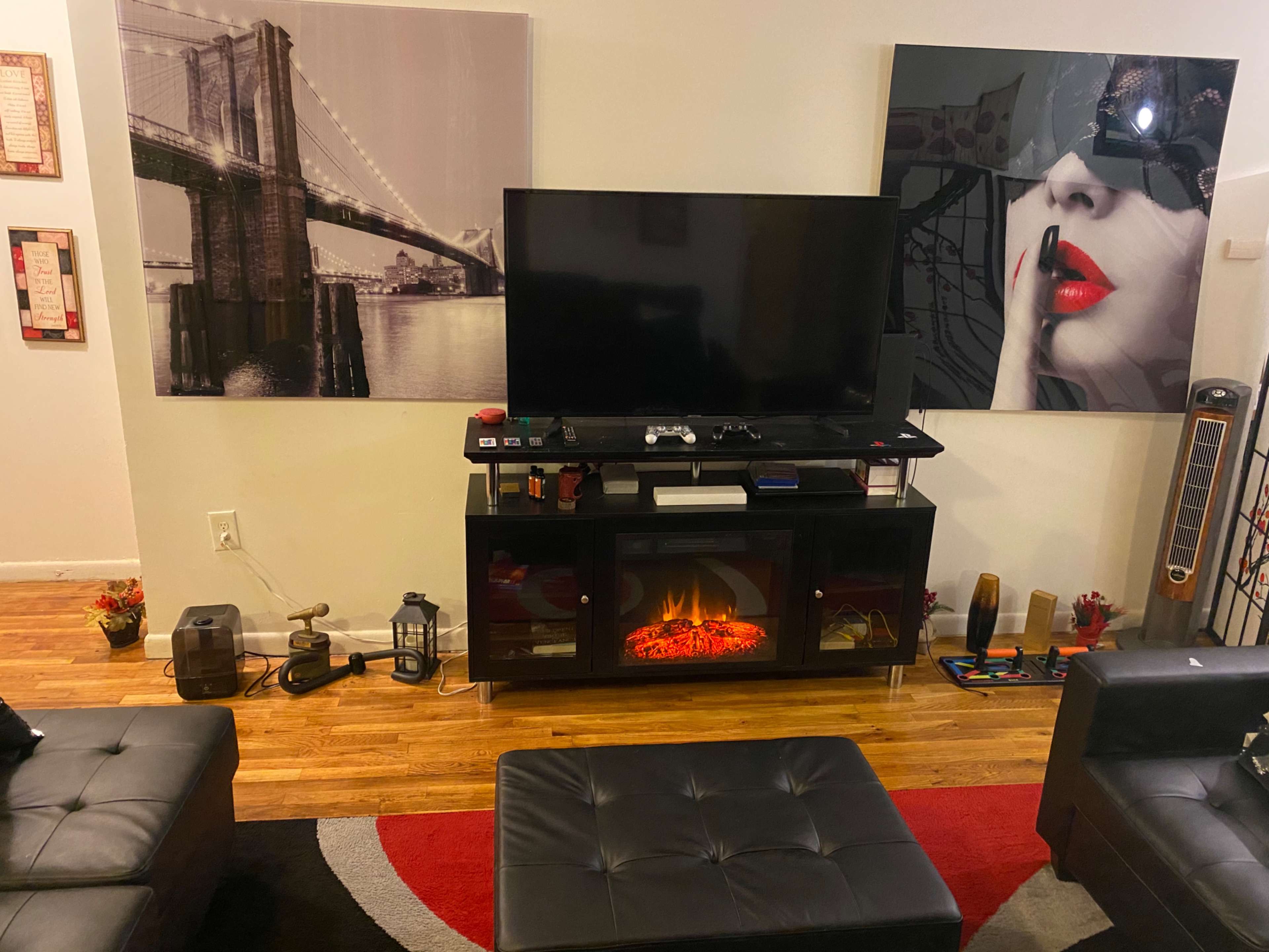 The image shows a living room with a television mounted on a stand in front of two large wall art pieces, a decorative fireplace, and black leather furniture arranged on a patterned rug.