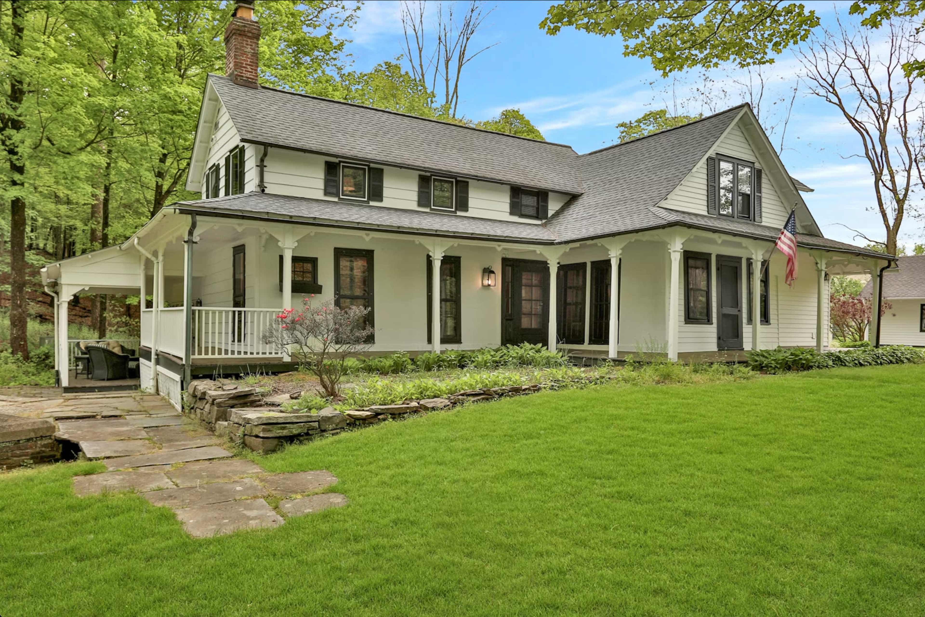 A white two-story house with a sloped roof is surrounded by green grass and trees, featuring a front porch and American flag.