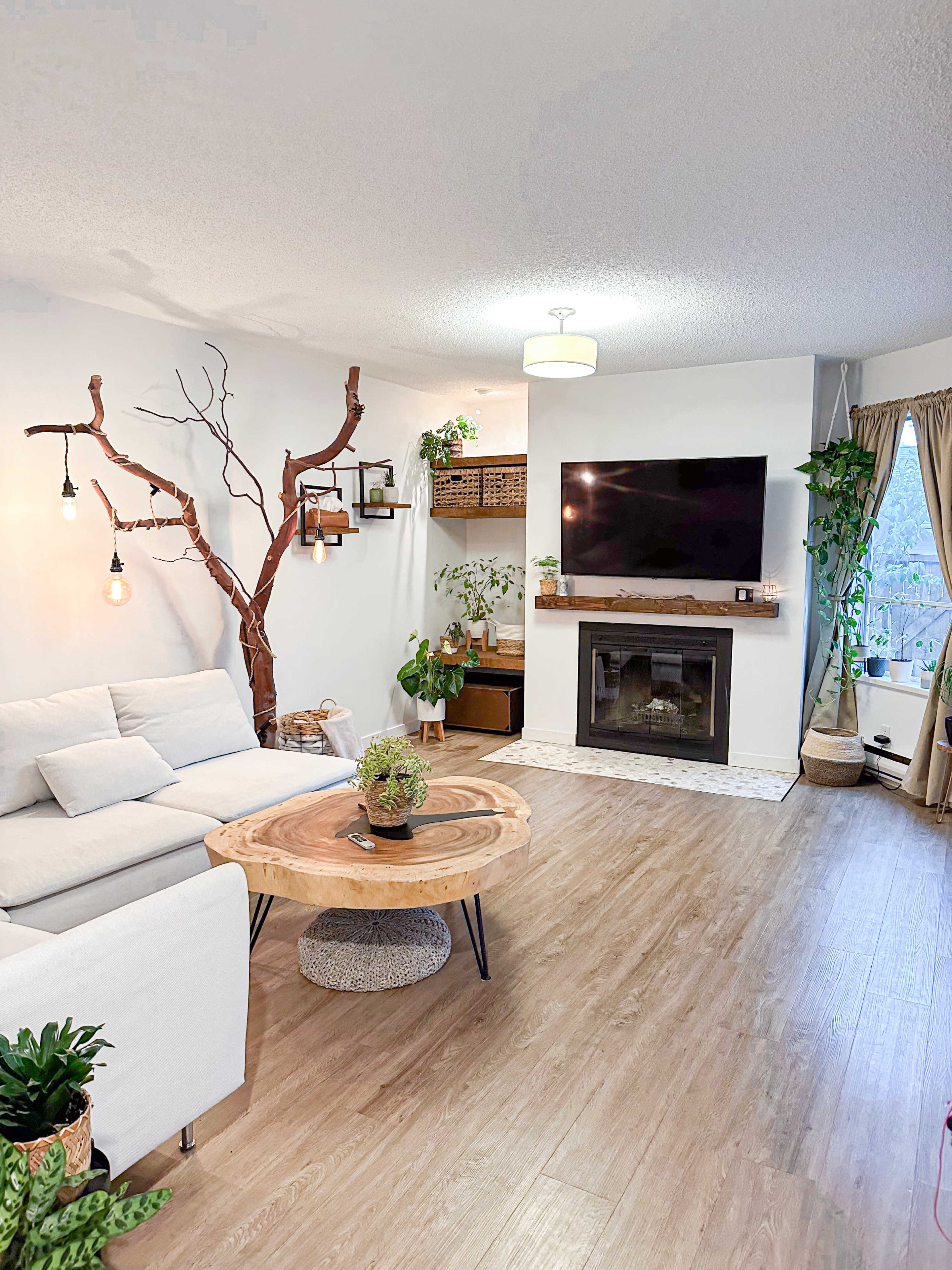 A modern living room featuring a light-colored couch, a wooden coffee table, a wall-mounted TV, and decorative plants, with a unique tree branch structure and hanging lights.