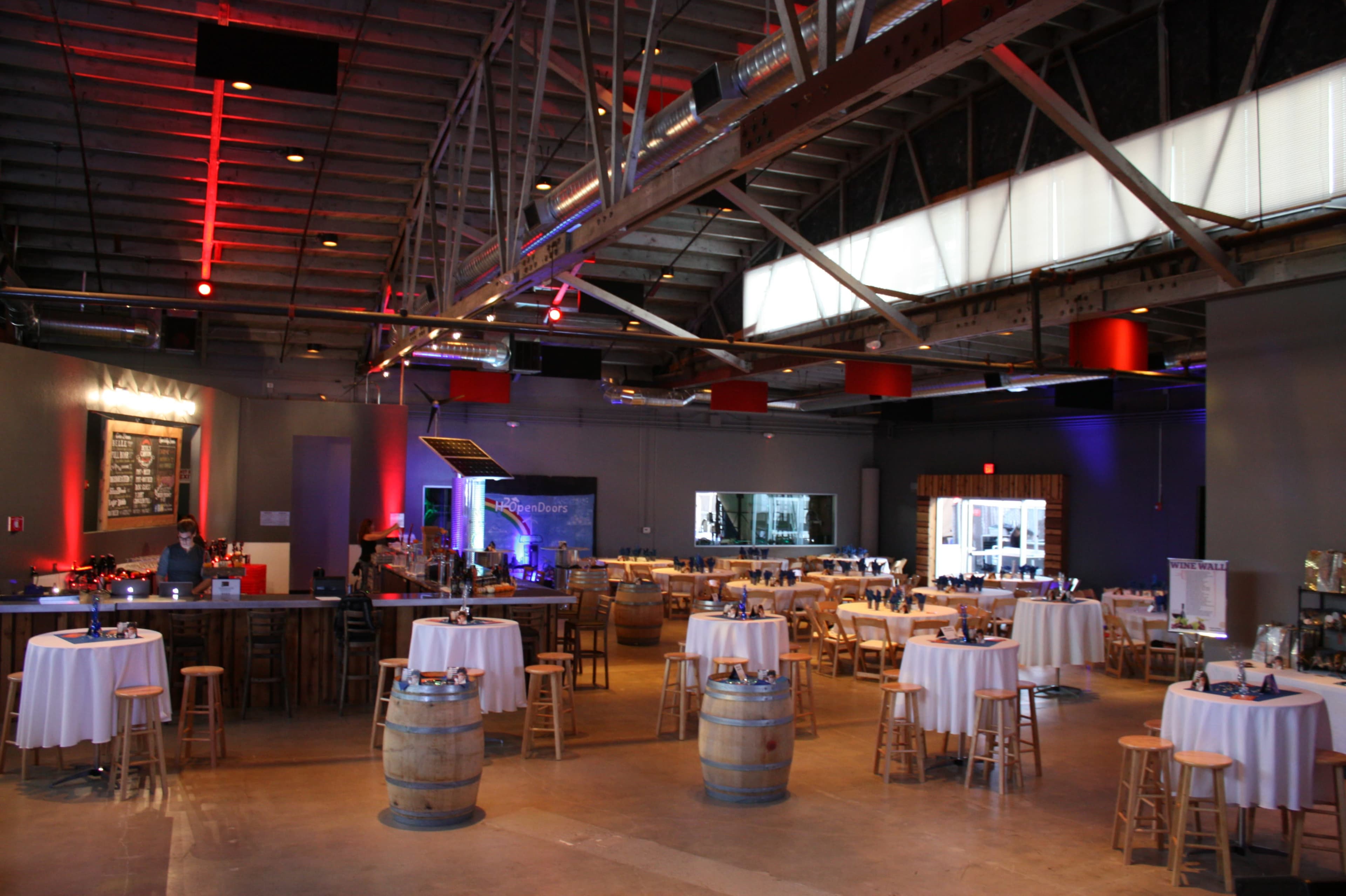 A spacious event venue with circular tables set for dining, surrounded by bar stools, and a bar area in the back.