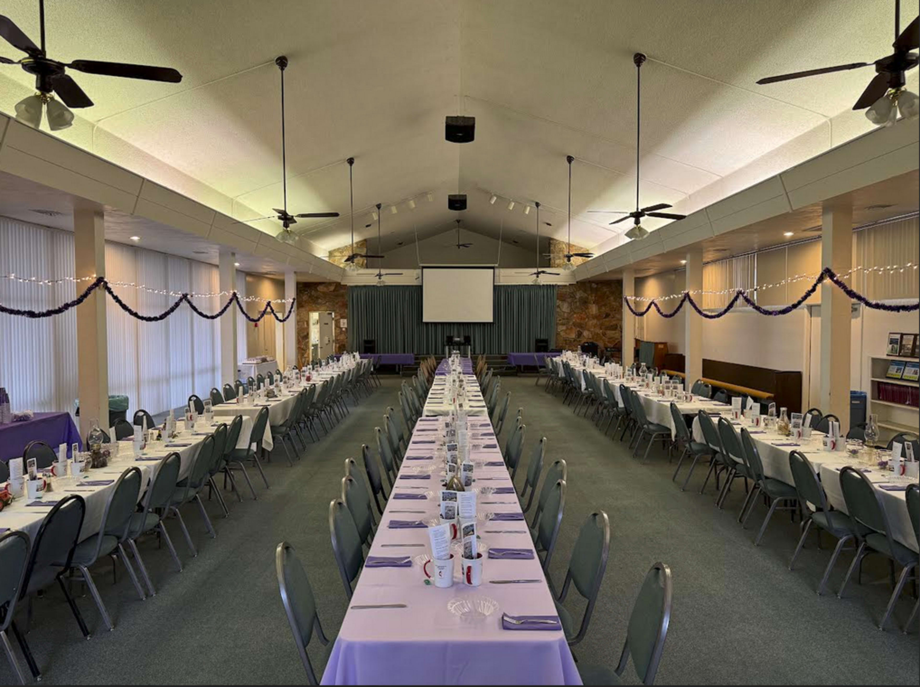 A long dining table is set up in an indoor hall, with chairs arranged on either side and decorations along the ceiling.