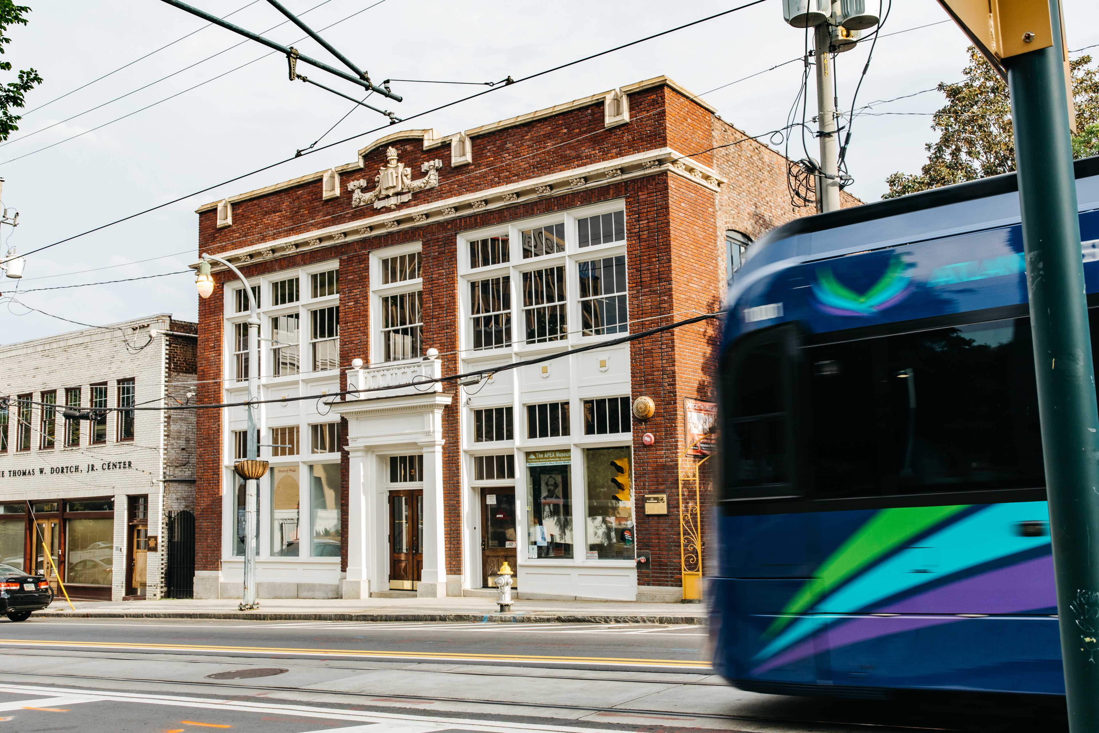 A historic brick building with large windows stands beside a busy street, while a streetcar passes by in motion.