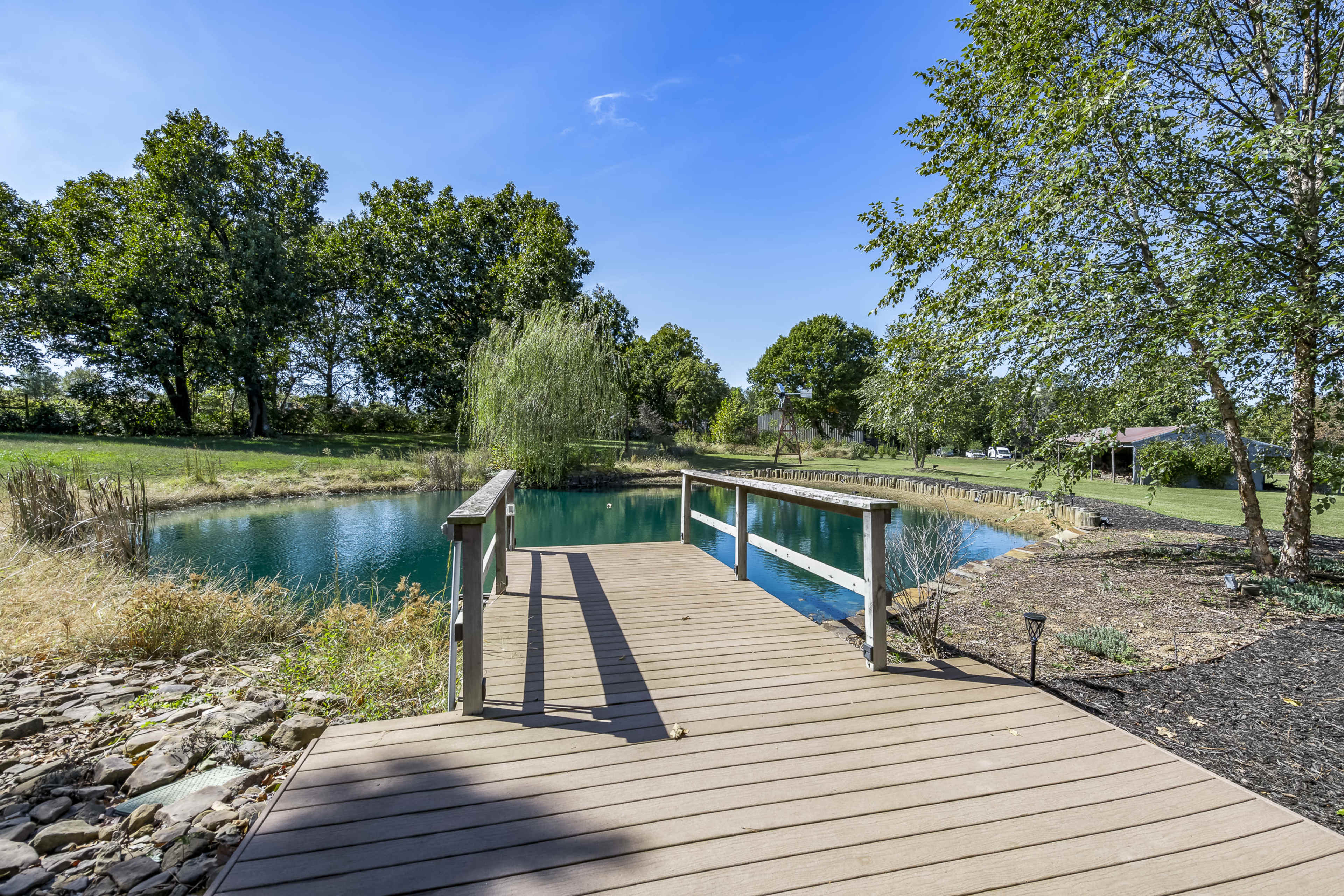 A wooden dock extends over a calm pond surrounded by trees and grassy areas under a clear blue sky.
