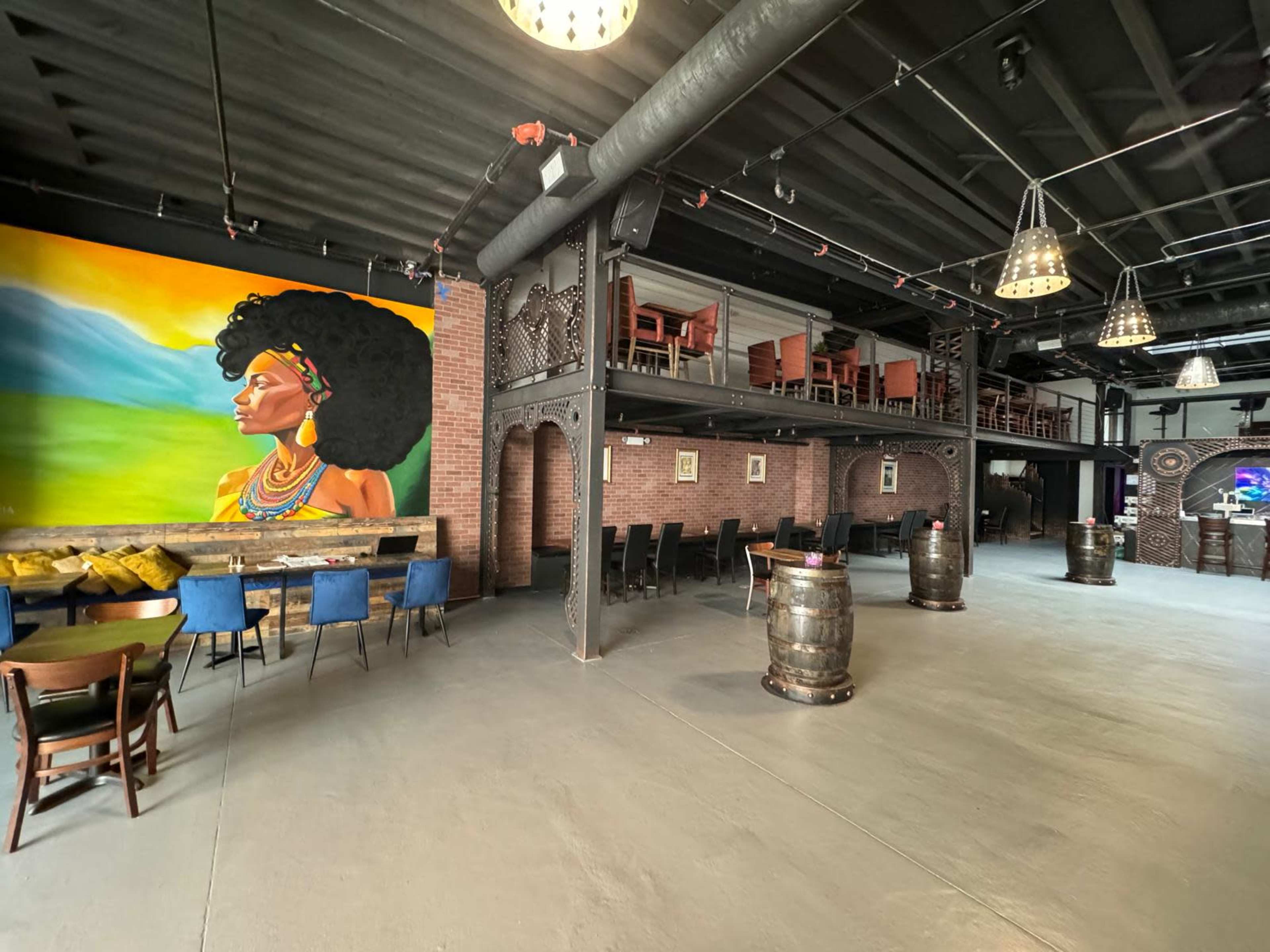 The image shows an interior space of a venue featuring a large colorful mural of a woman with an afro on one wall, wooden tables and chairs, and barrel-style seating arrangements.