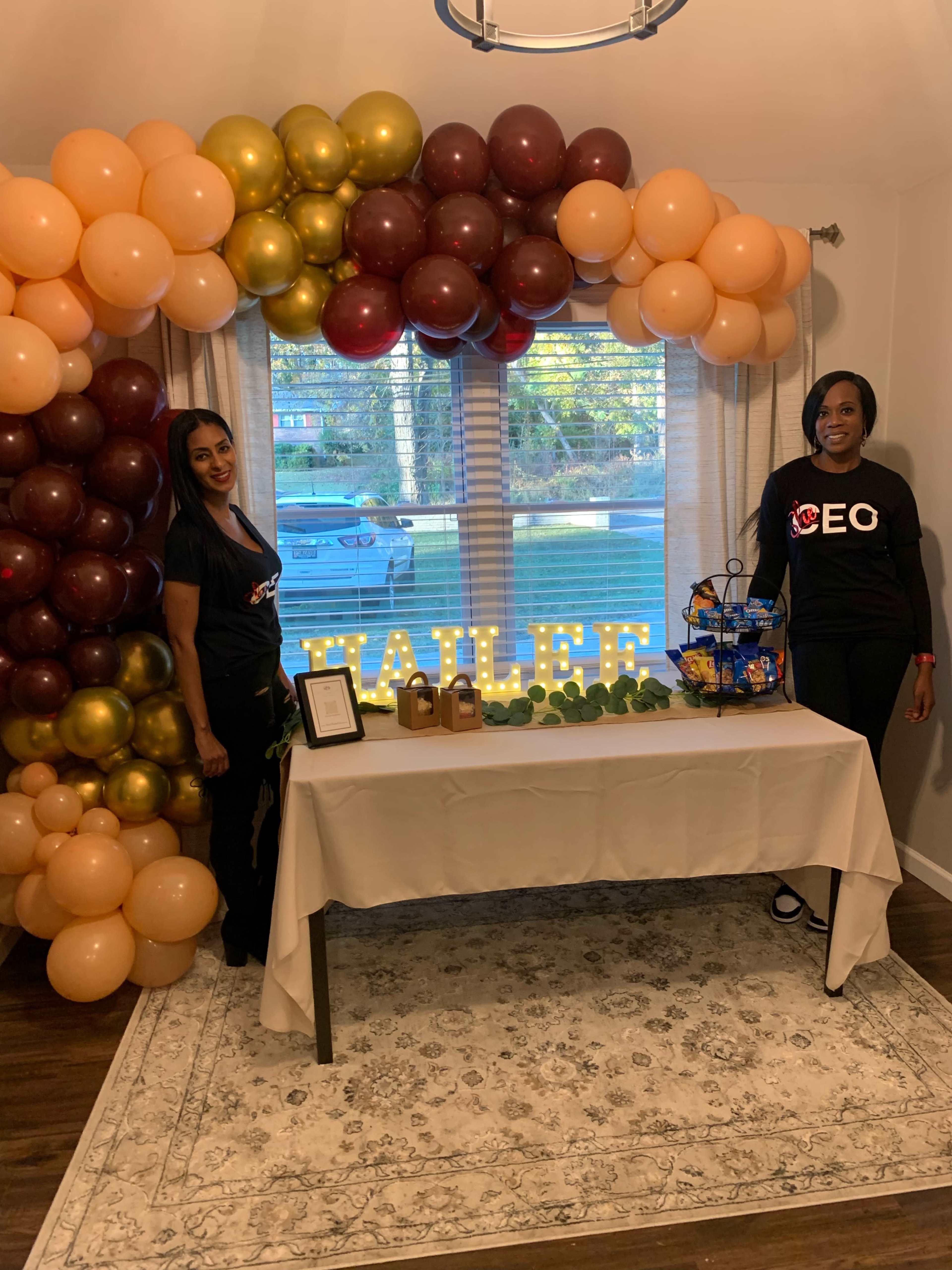 Two women stand beside a decorated table under a balloon arch, with the name "HAILEE" displayed in lights.