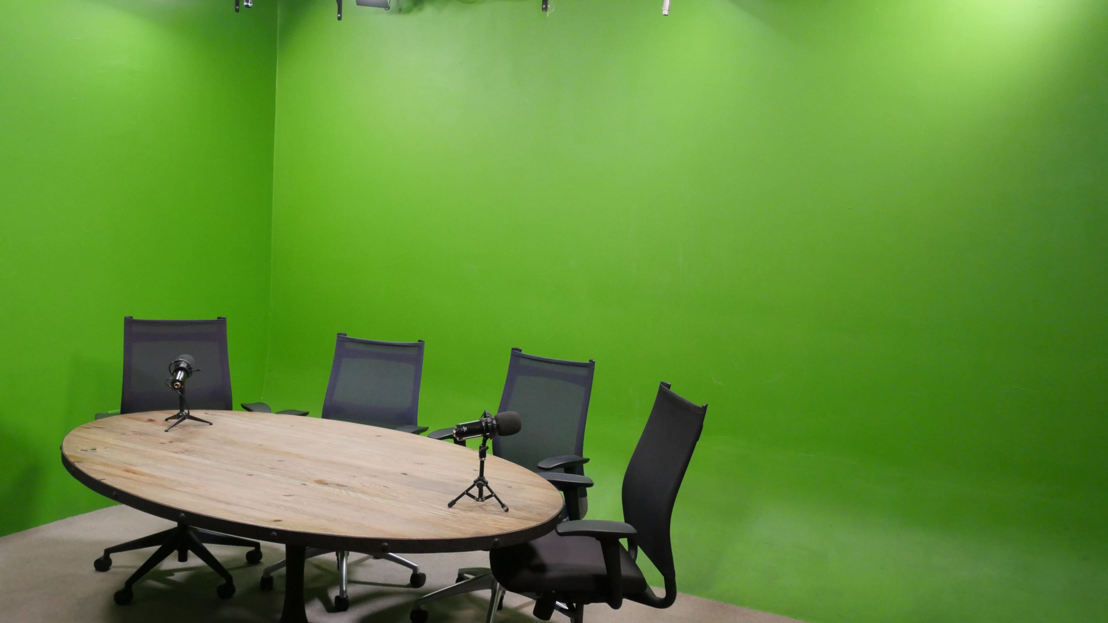 A circular wooden table with five chairs is placed in a room with green walls, and two microphones are positioned on the table.