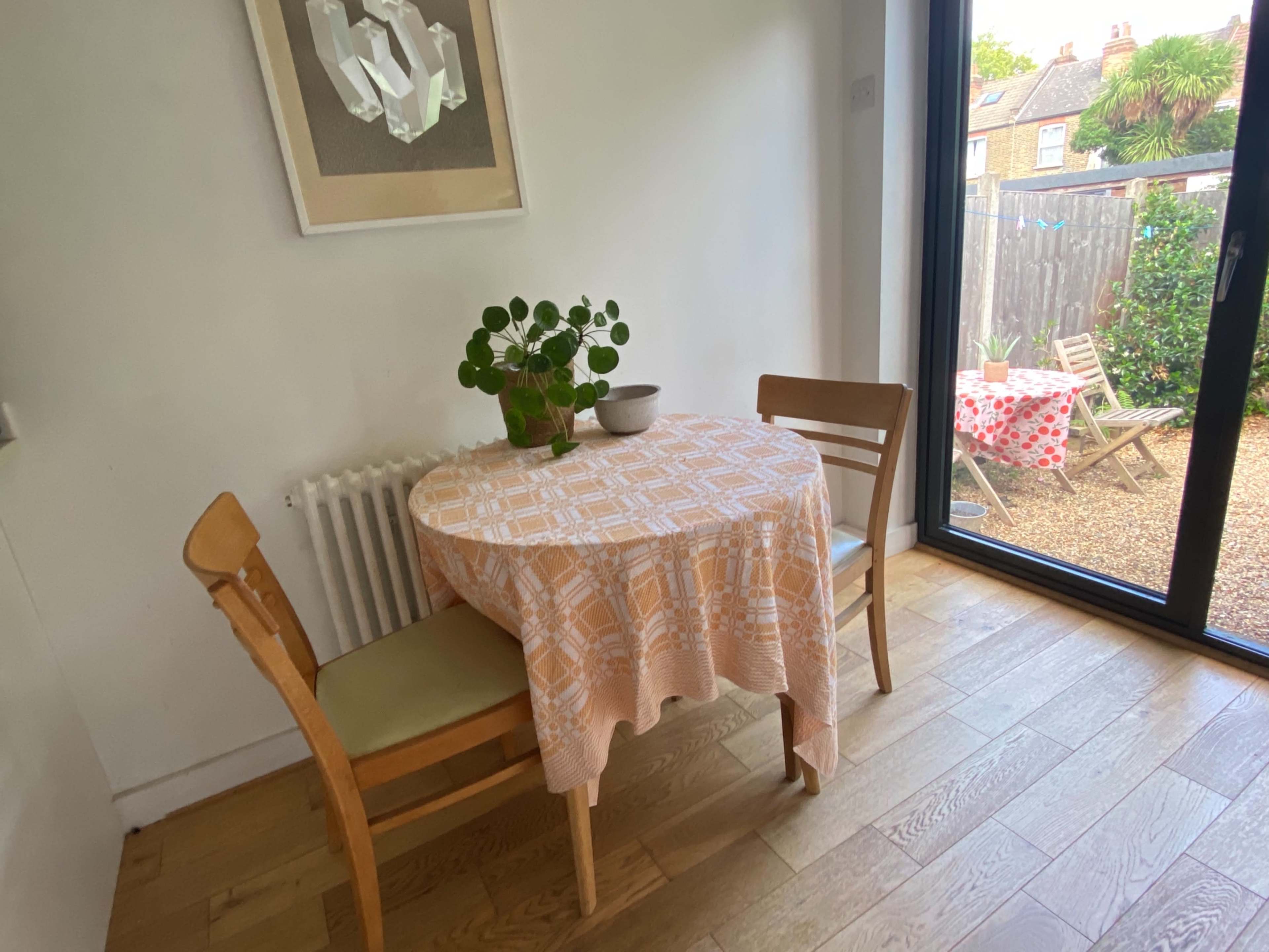 A small dining table with a tablecloth and a plant centerpiece is positioned near a large glass door leading to an outdoor area, with a second table visible in the background.