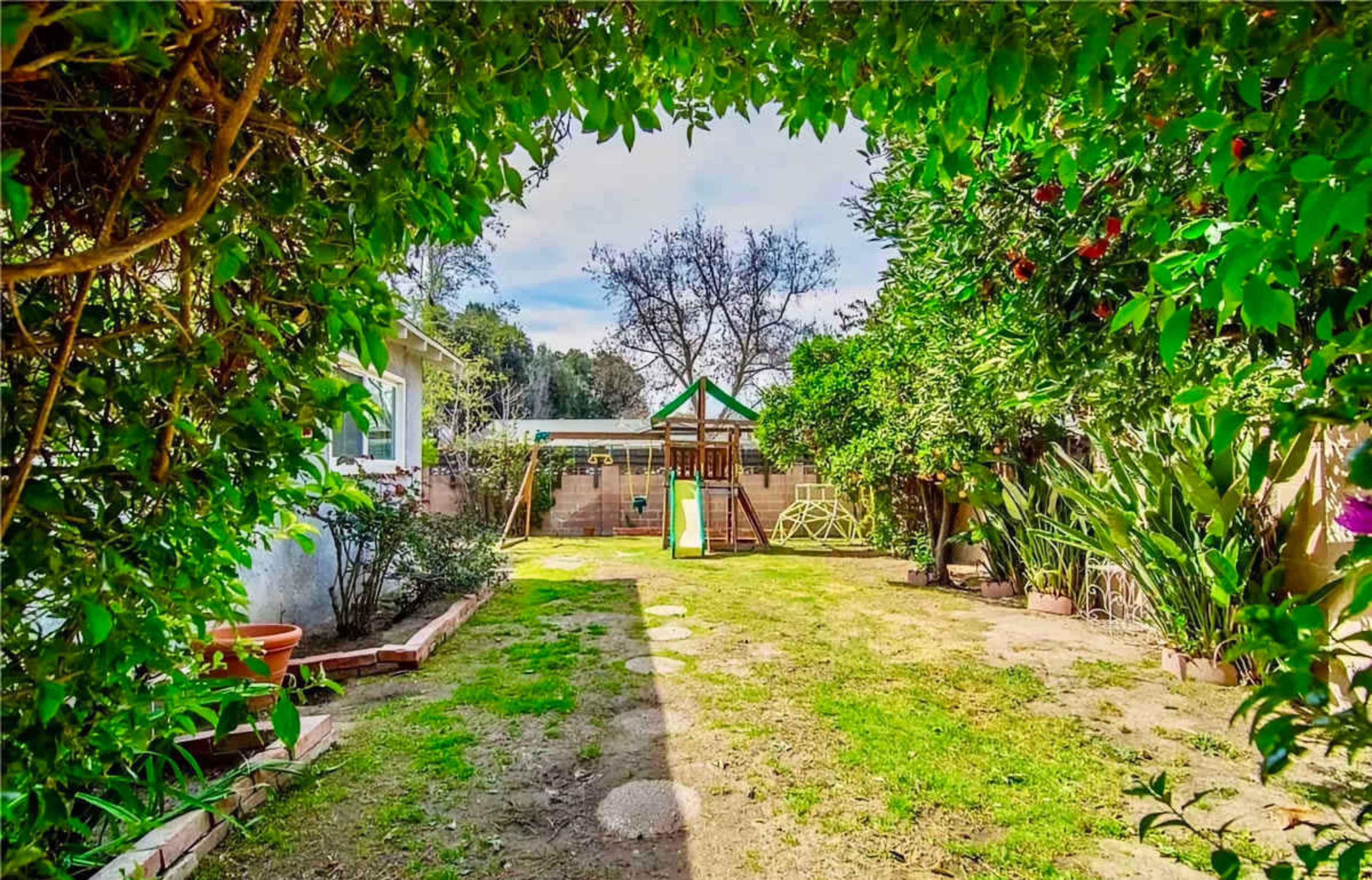 The image shows a backyard with a grassy area, a children’s playset, and various trees and shrubs.
