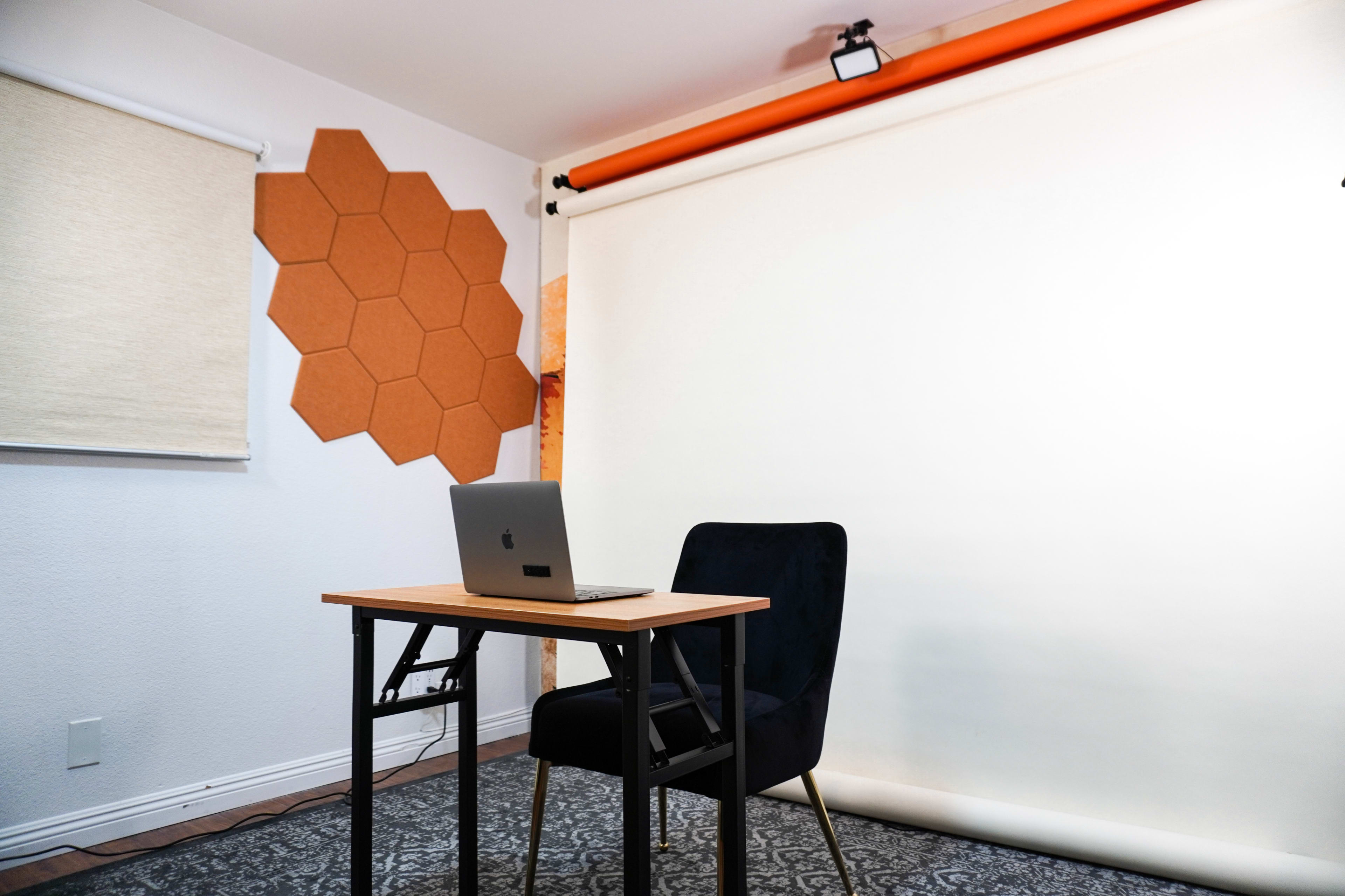 A simple workspace with a black chair and a laptop positioned on a wooden table, alongside a light-colored wall with hexagonal panels and a rolled-up backdrop.