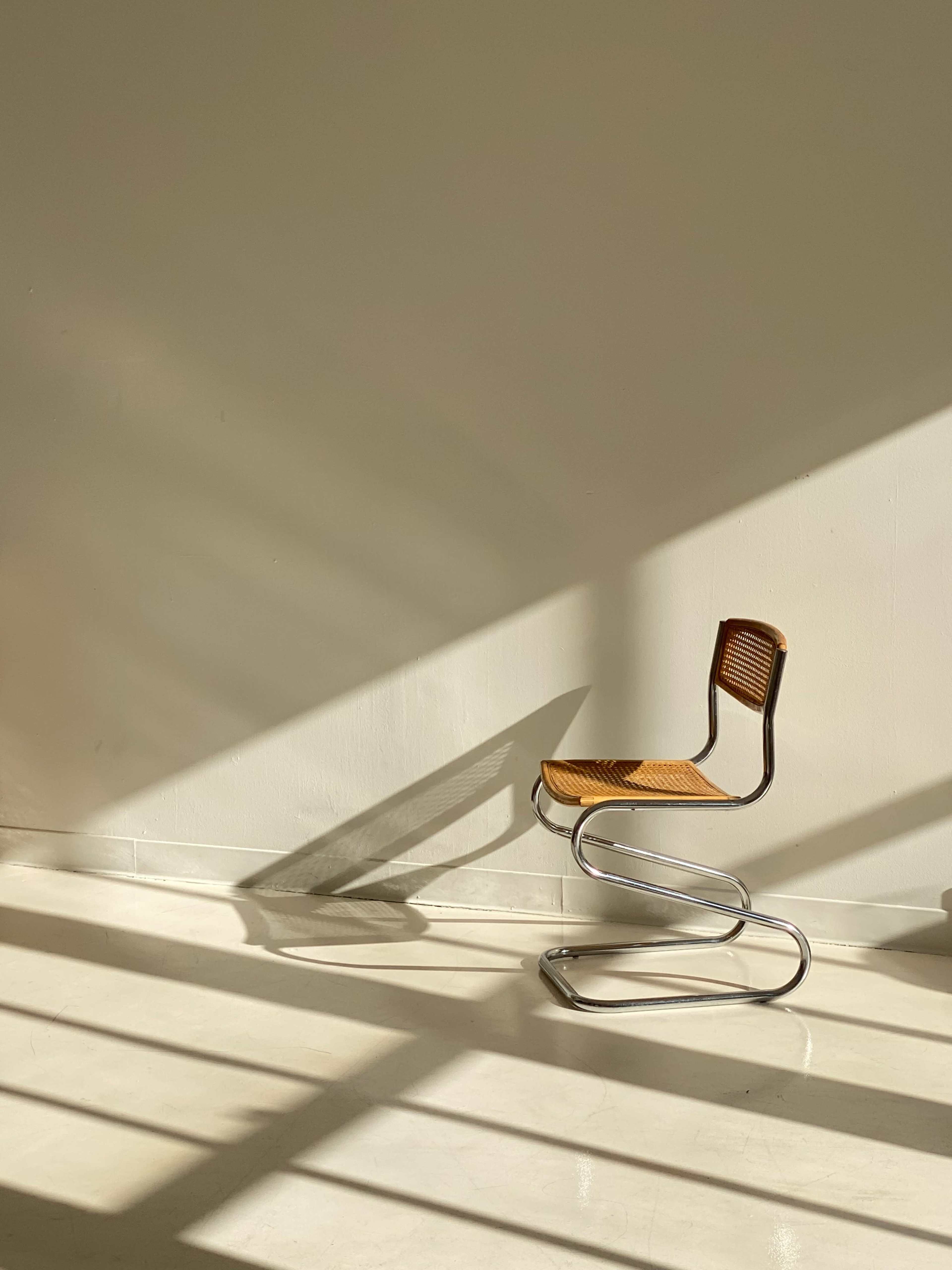 A modern metal chair with a woven seat is positioned against a blank wall, casting shadows from sunlight filtering through nearby blinds.
