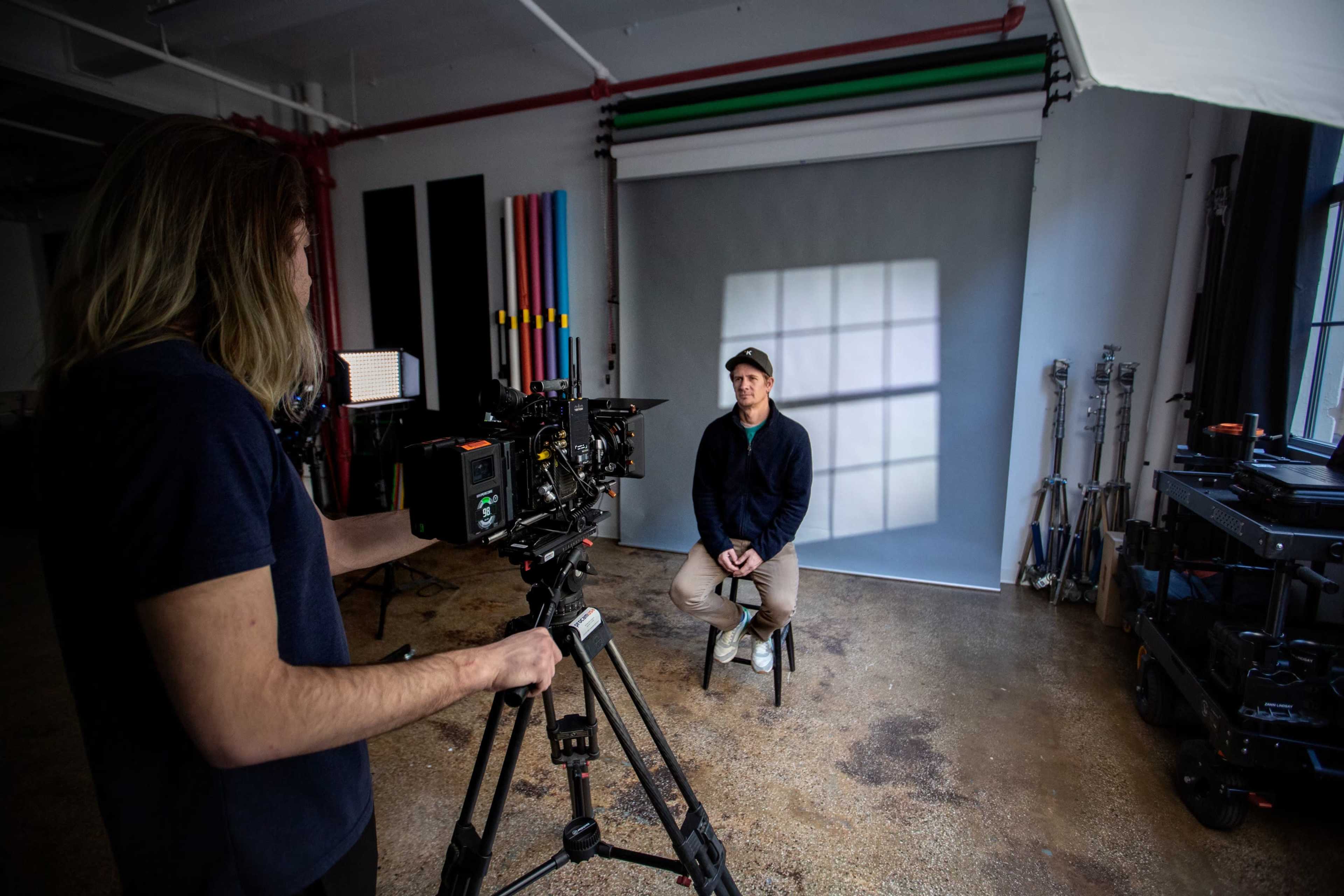 A man is seated on a chair in a studio while another person operates a camera directed at him.
