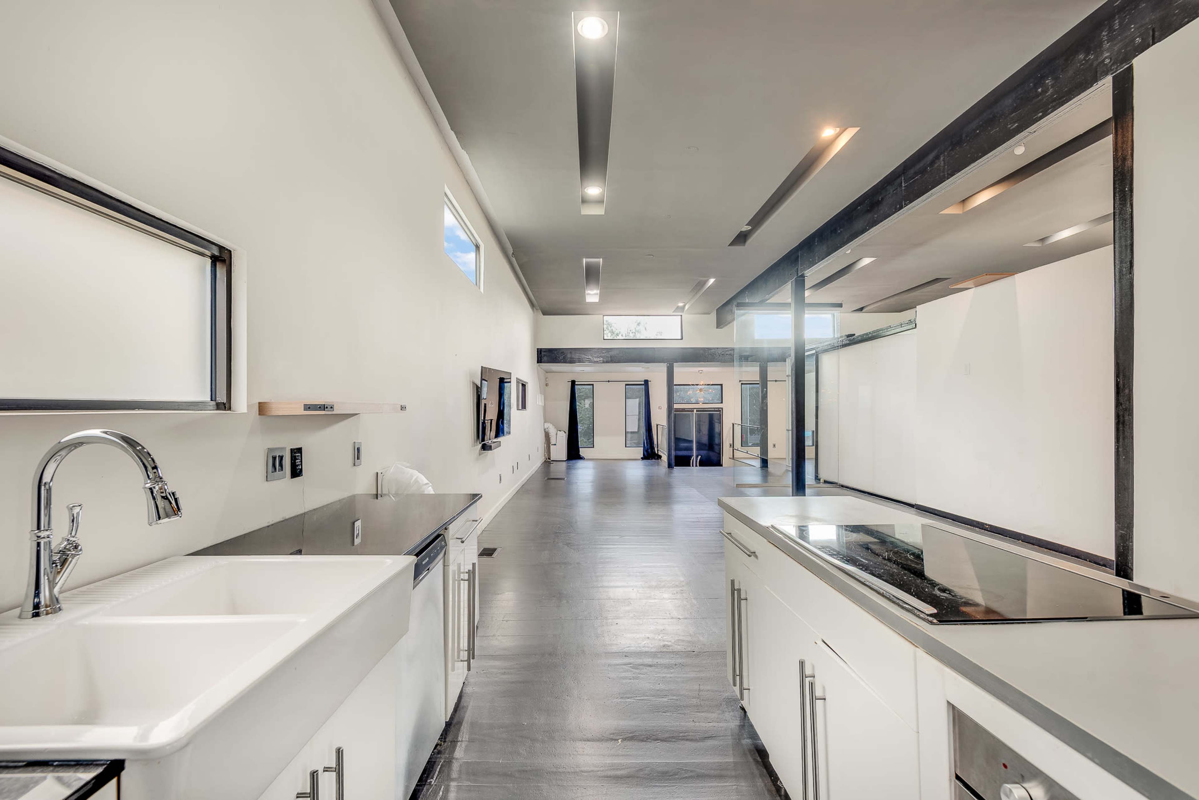 The image shows a modern, minimalist kitchen and dining area with white cabinetry, a large sink, and an open layout leading to a bright, spacious hallway.