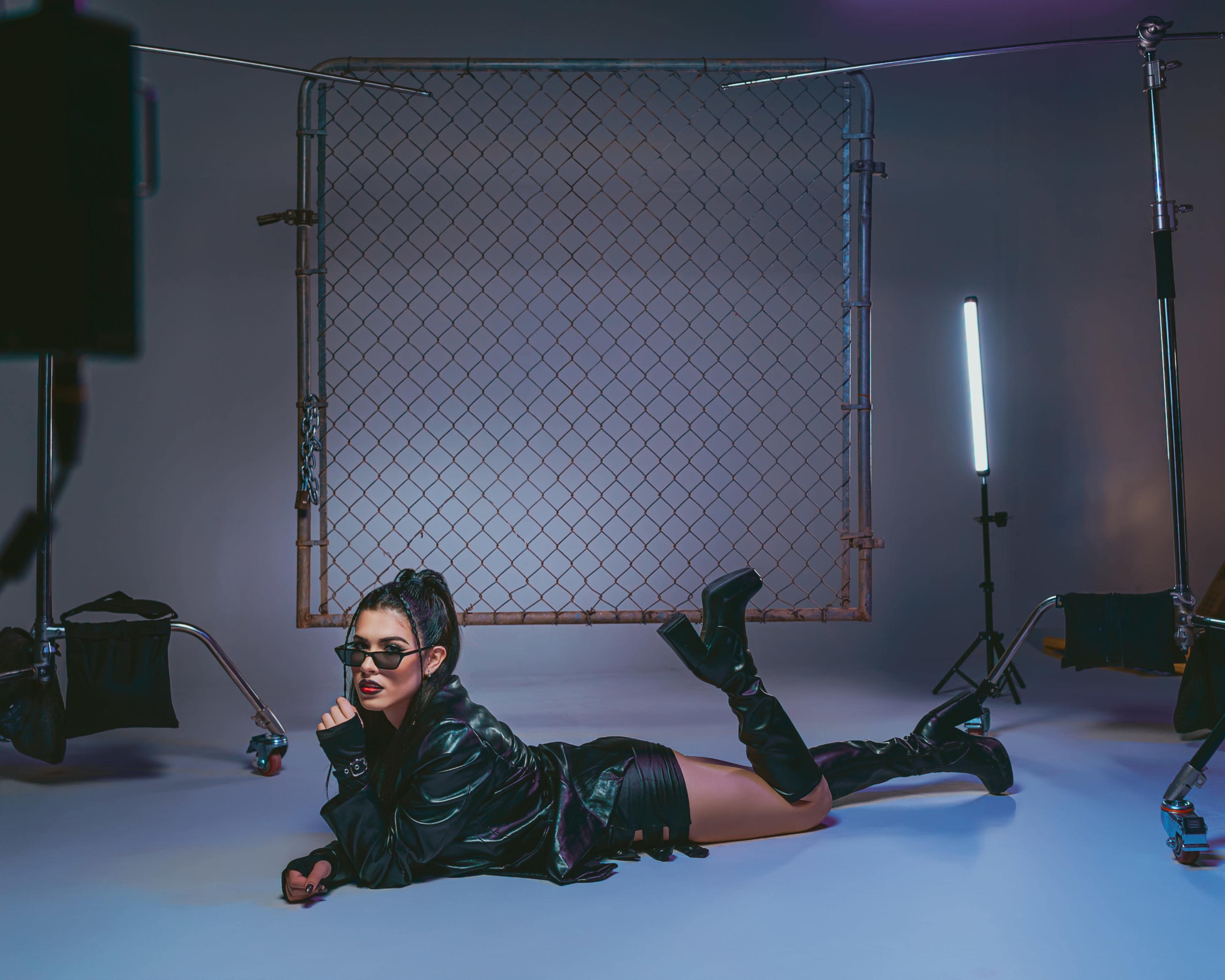 A model poses on the floor in a dark jacket and sunglasses, with a chain-link backdrop and studio lights illuminating the scene.