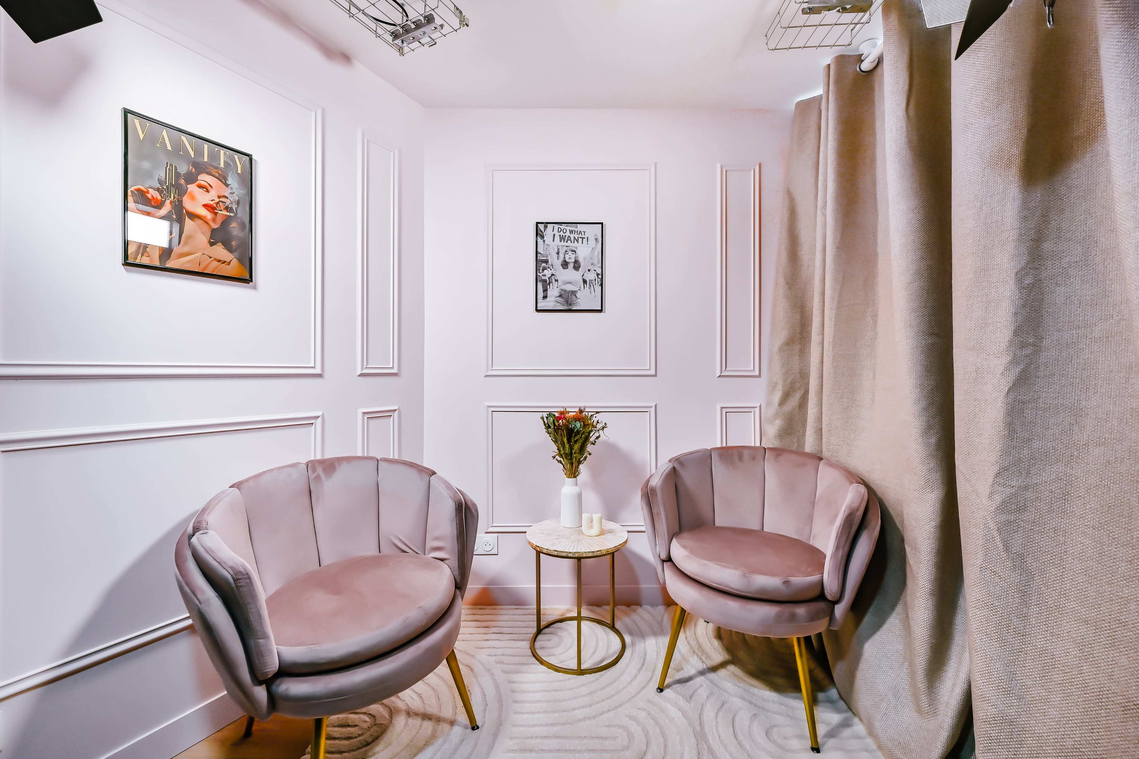 The image shows a small, elegantly designed seating area with two plush chairs, a round table, and framed artwork on the walls.