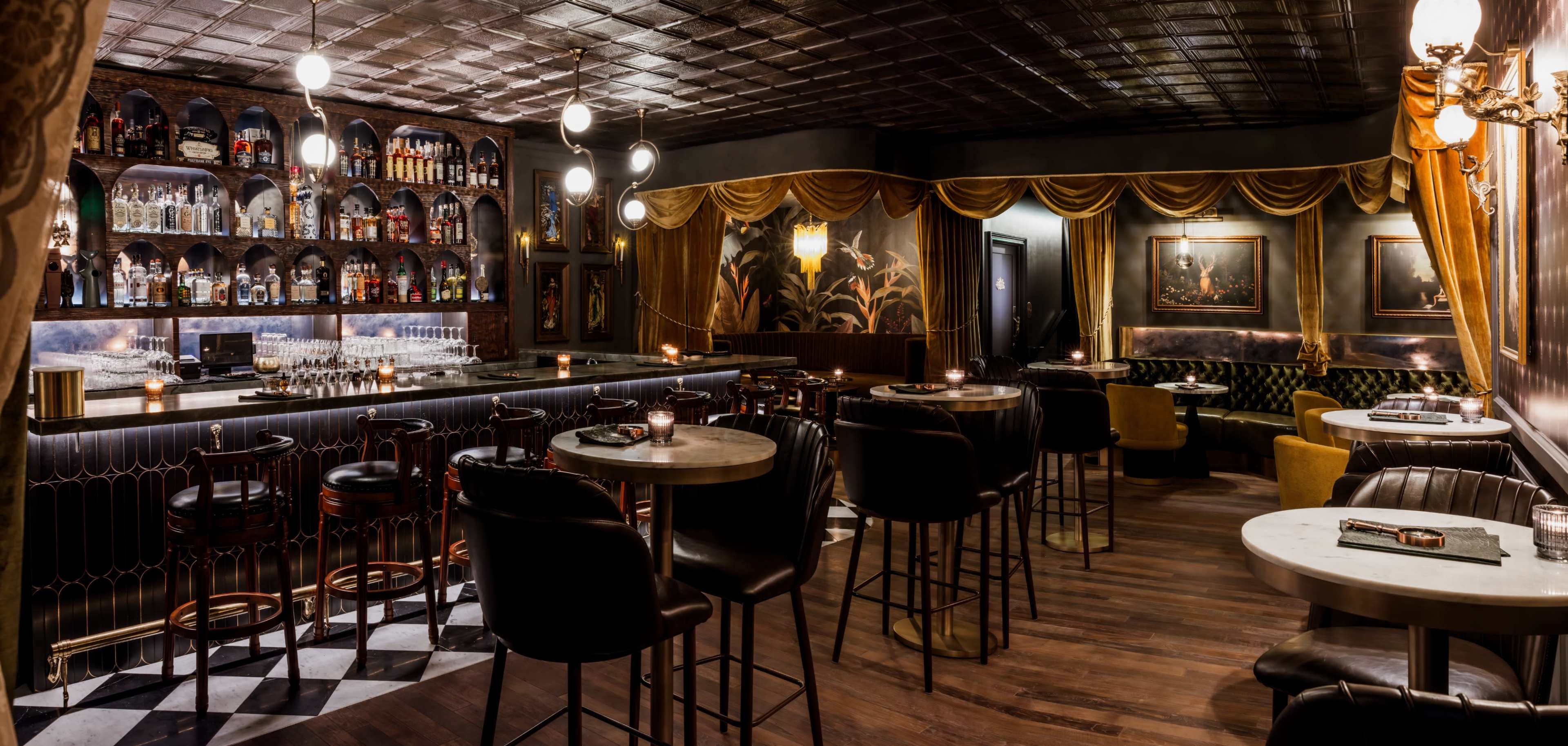 The image shows a dimly lit bar with a mirrored back shelf displaying various bottles, elegant seating, and decorative wall accents.
