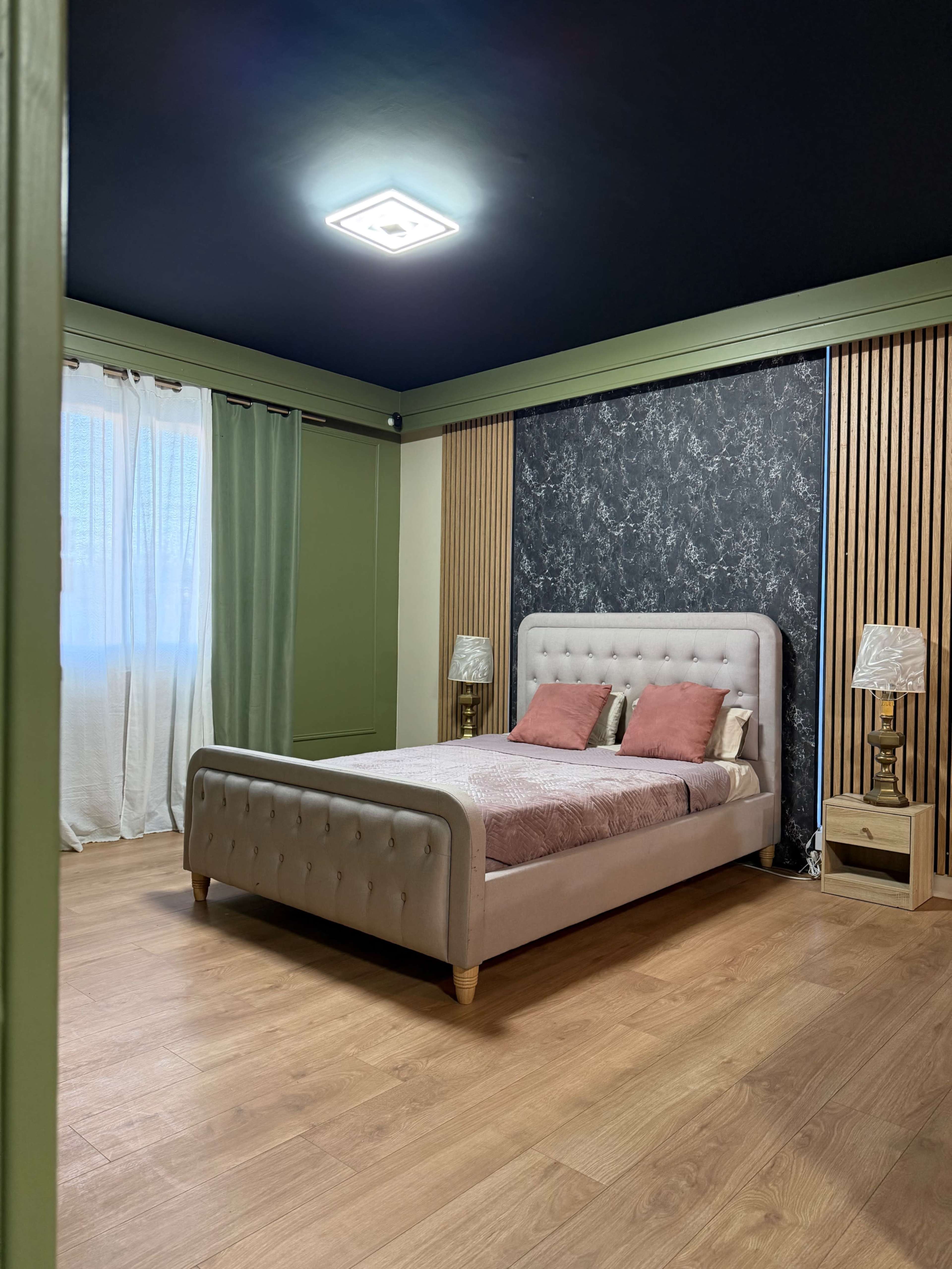 The image shows a modern bedroom featuring a beige upholstered bed with pink pillows, wooden flooring, green walls, and a dark accent wall behind the bed.