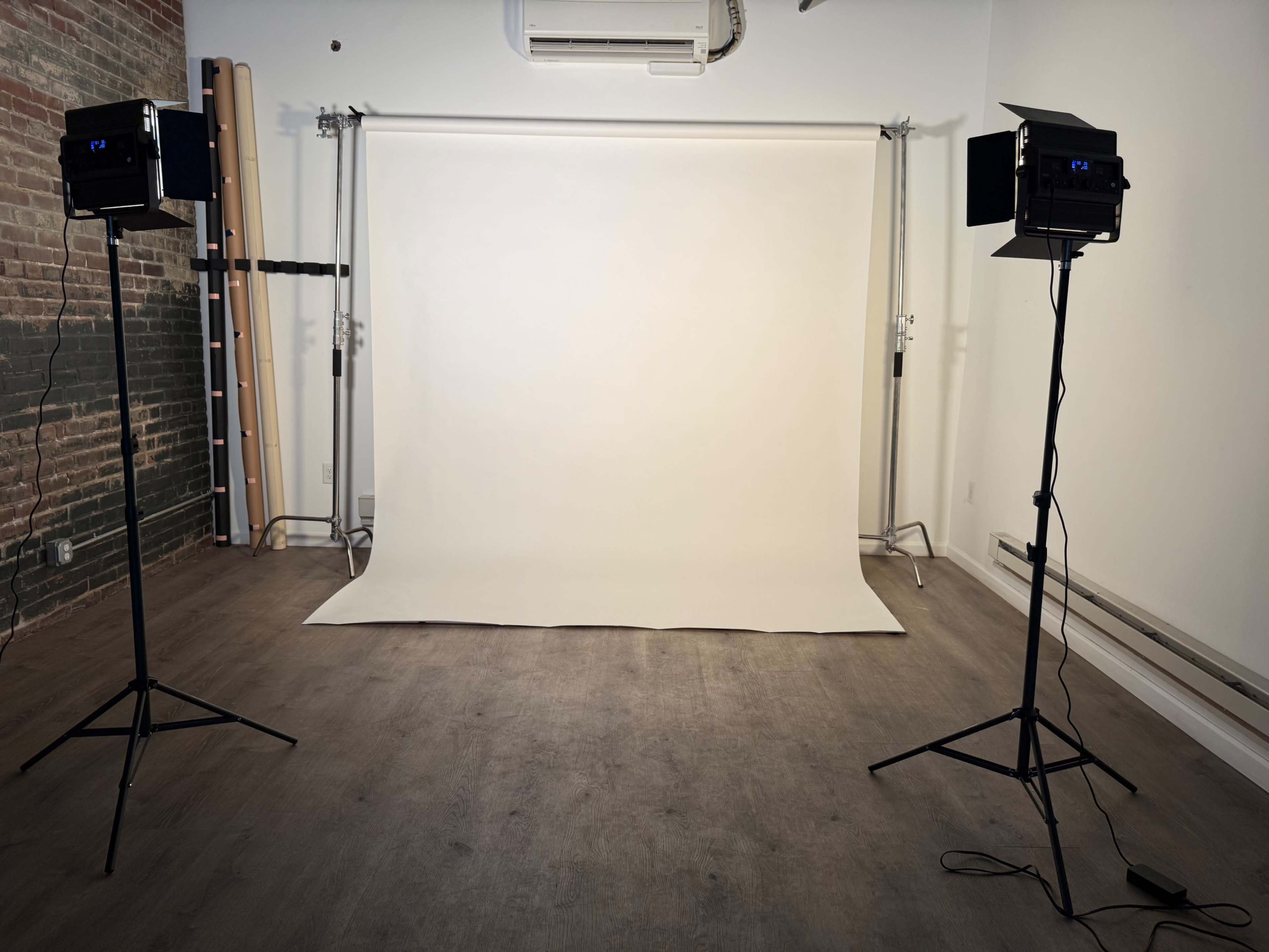 The image shows a photography studio setup with two lights on stands facing a plain white backdrop in a room with exposed brick walls and wooden flooring.