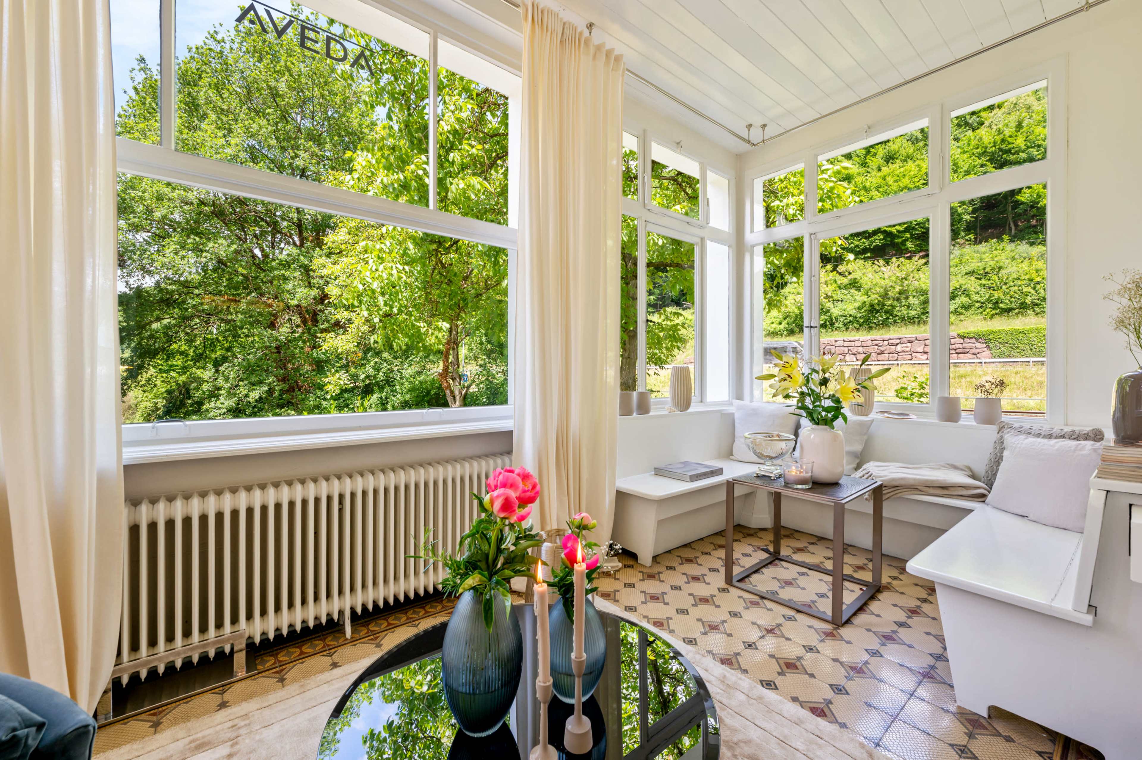 The image shows a bright sunroom with large windows surrounded by greenery, featuring a coffee table, decorative vases, and a radiator.