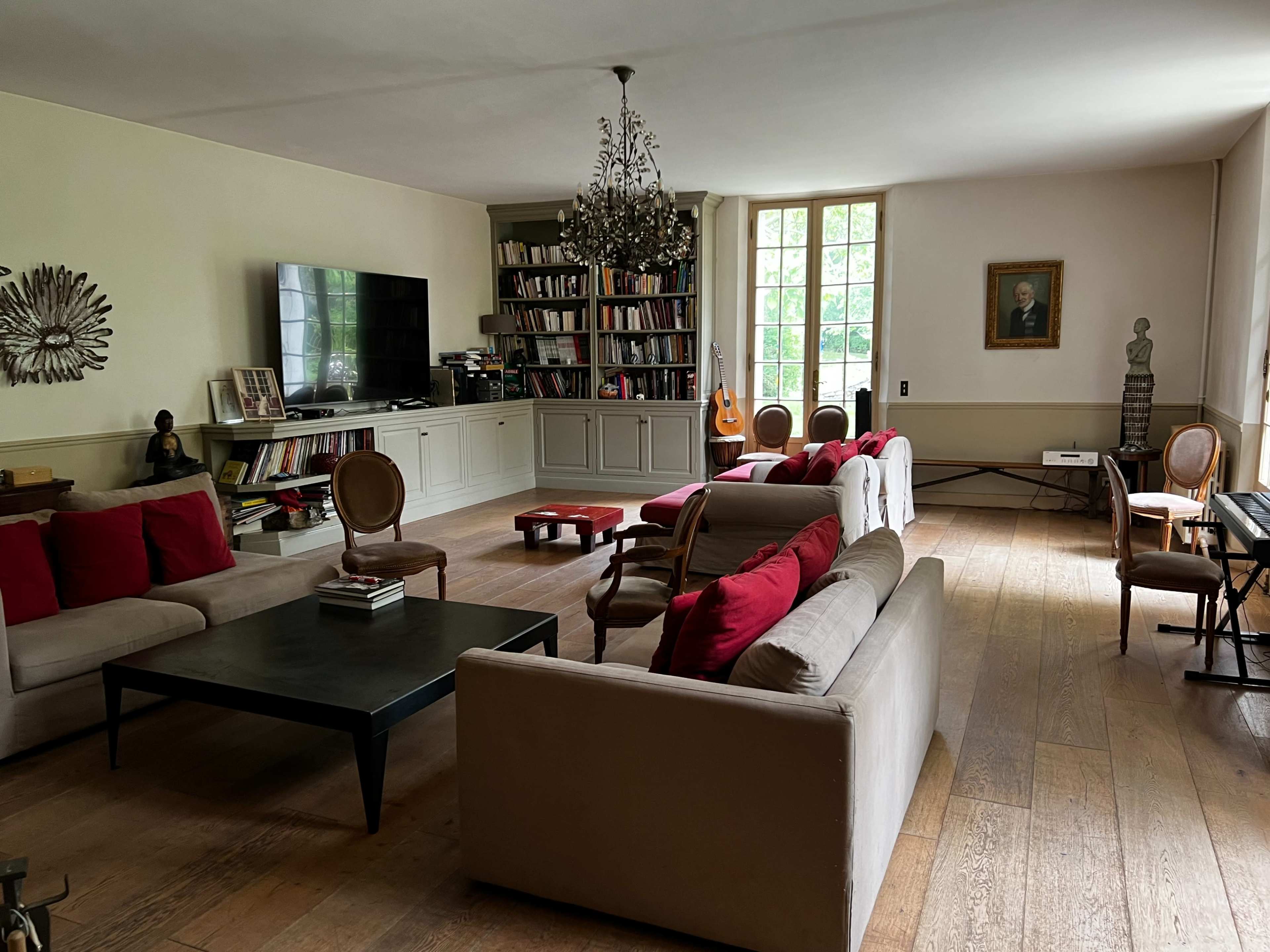 The room features a spacious living area with multiple seating arrangements, a television, bookshelves, and a large window allowing natural light.