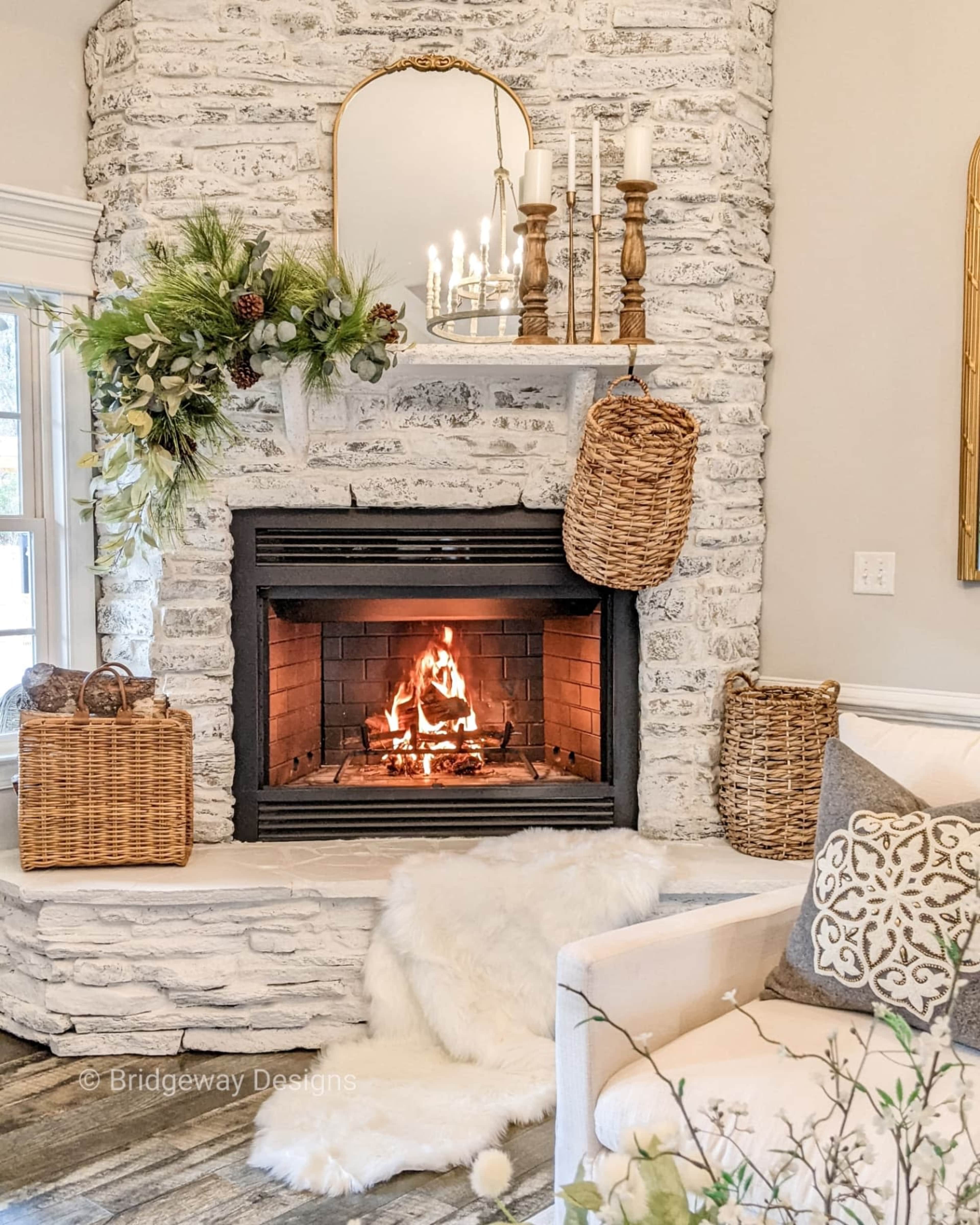 A cozy living room featuring a stone fireplace with a lit fire, surrounded by decorative elements like baskets and greenery, and a soft throw draped on a light sofa.