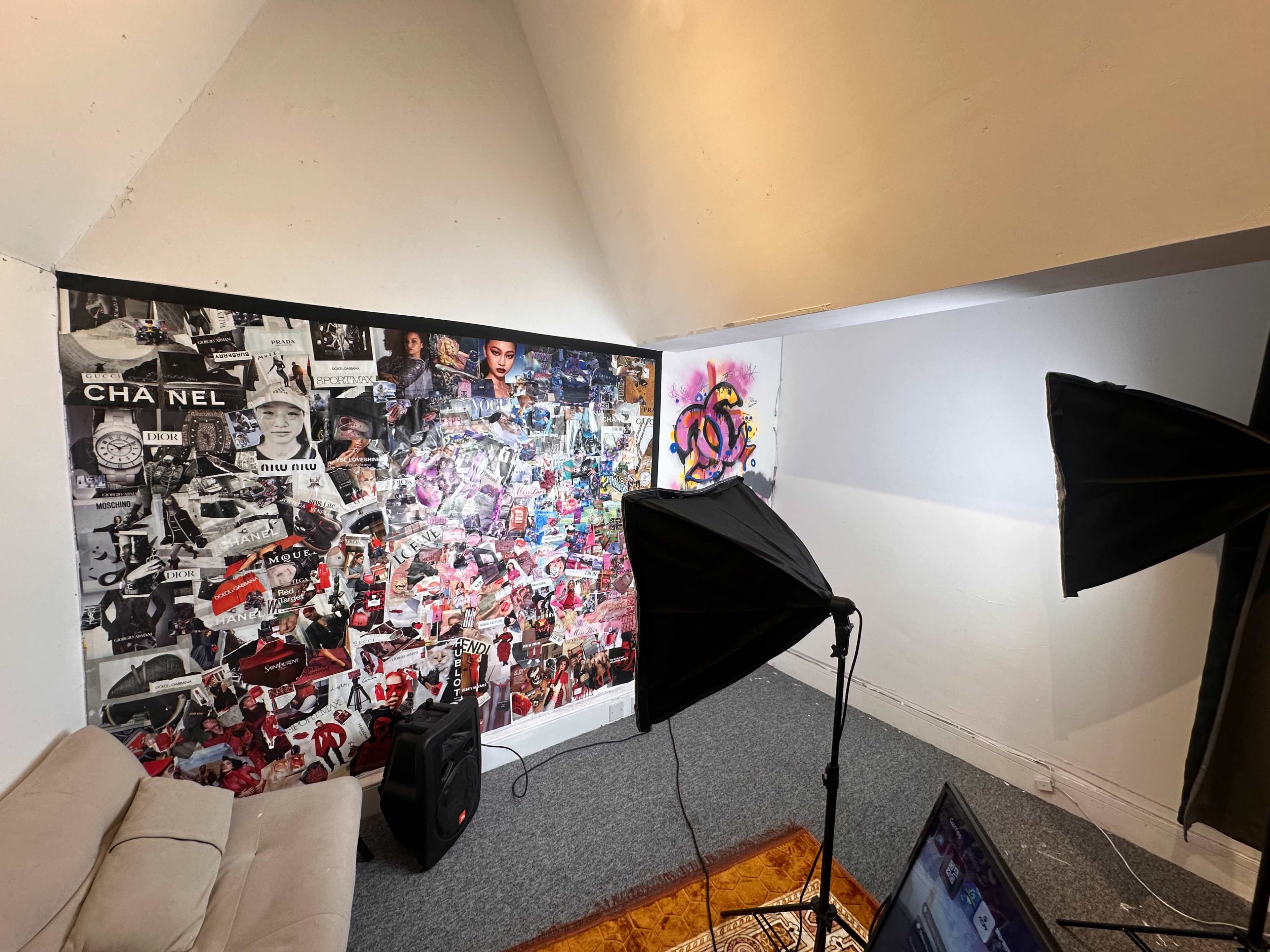 The image shows a small room with a collage of images and logos on one wall, two softbox lights set up, and a laptop on the floor.