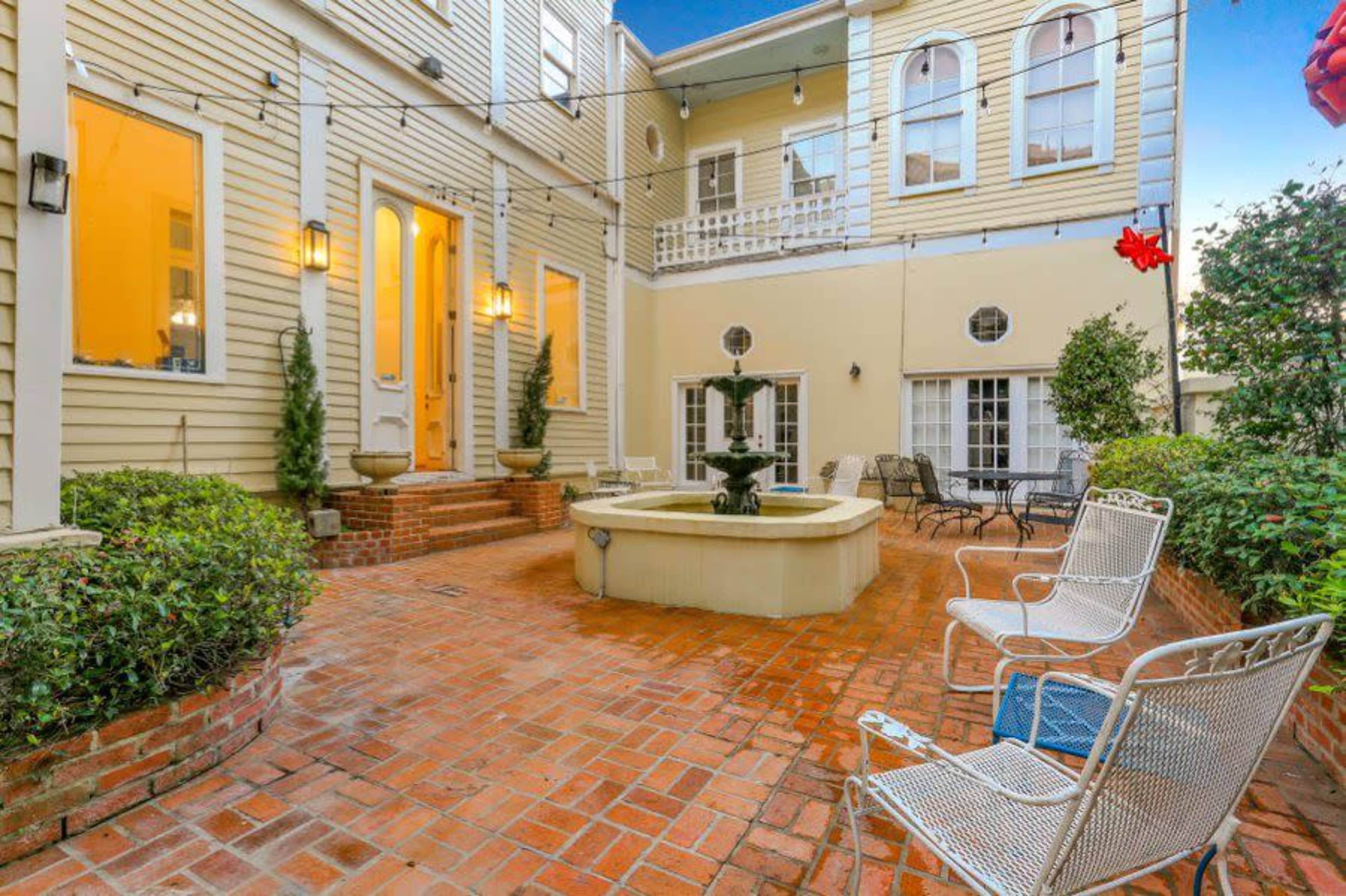 The image shows a brick patio area with a central fountain, surrounded by potted plants, chairs, and the exterior of a multi-story building.
