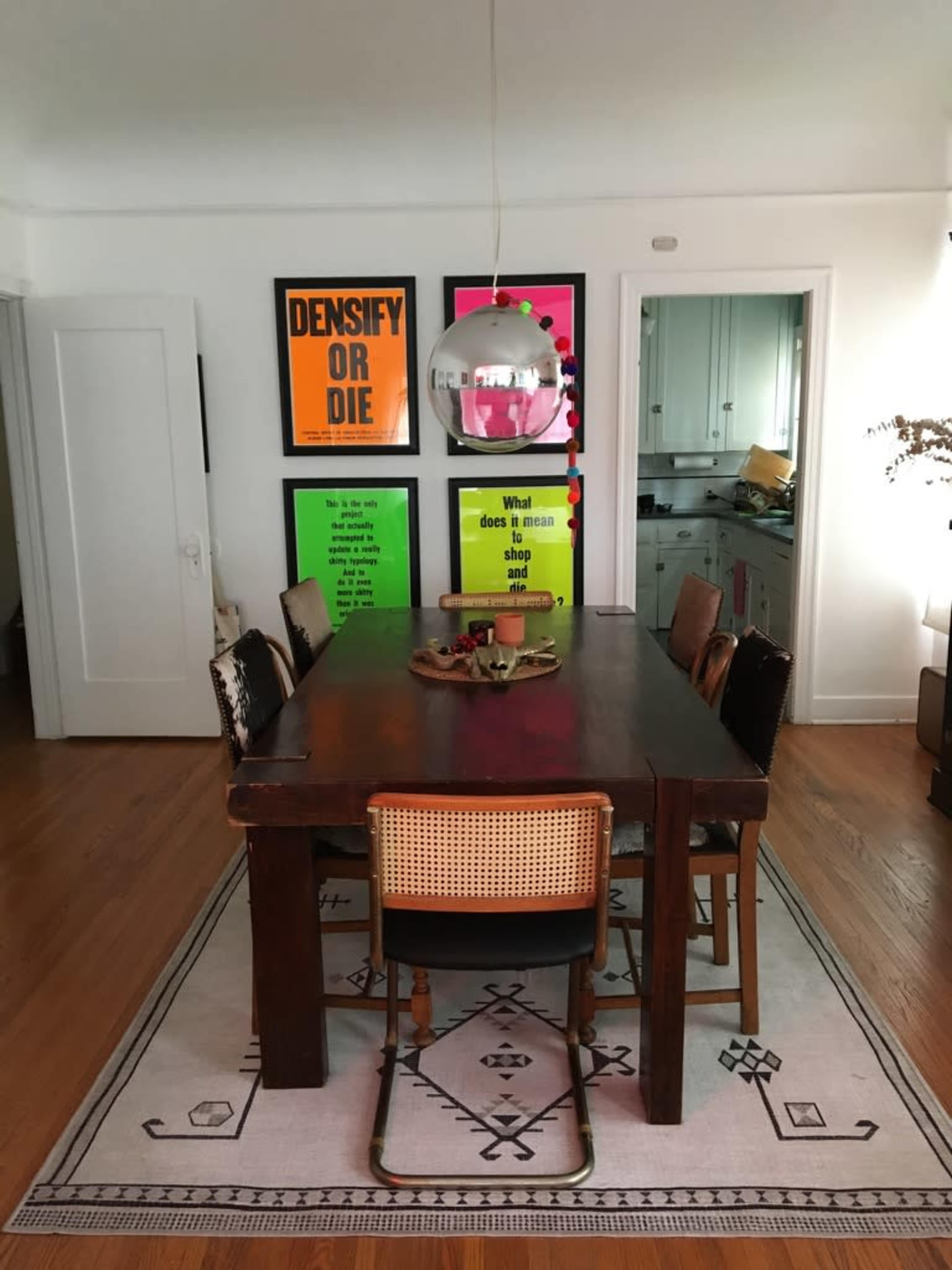 A wooden dining table with eight chairs is centered in a brightly lit room, featuring colorful framed posters on the wall and a modern pendant light hanging above.