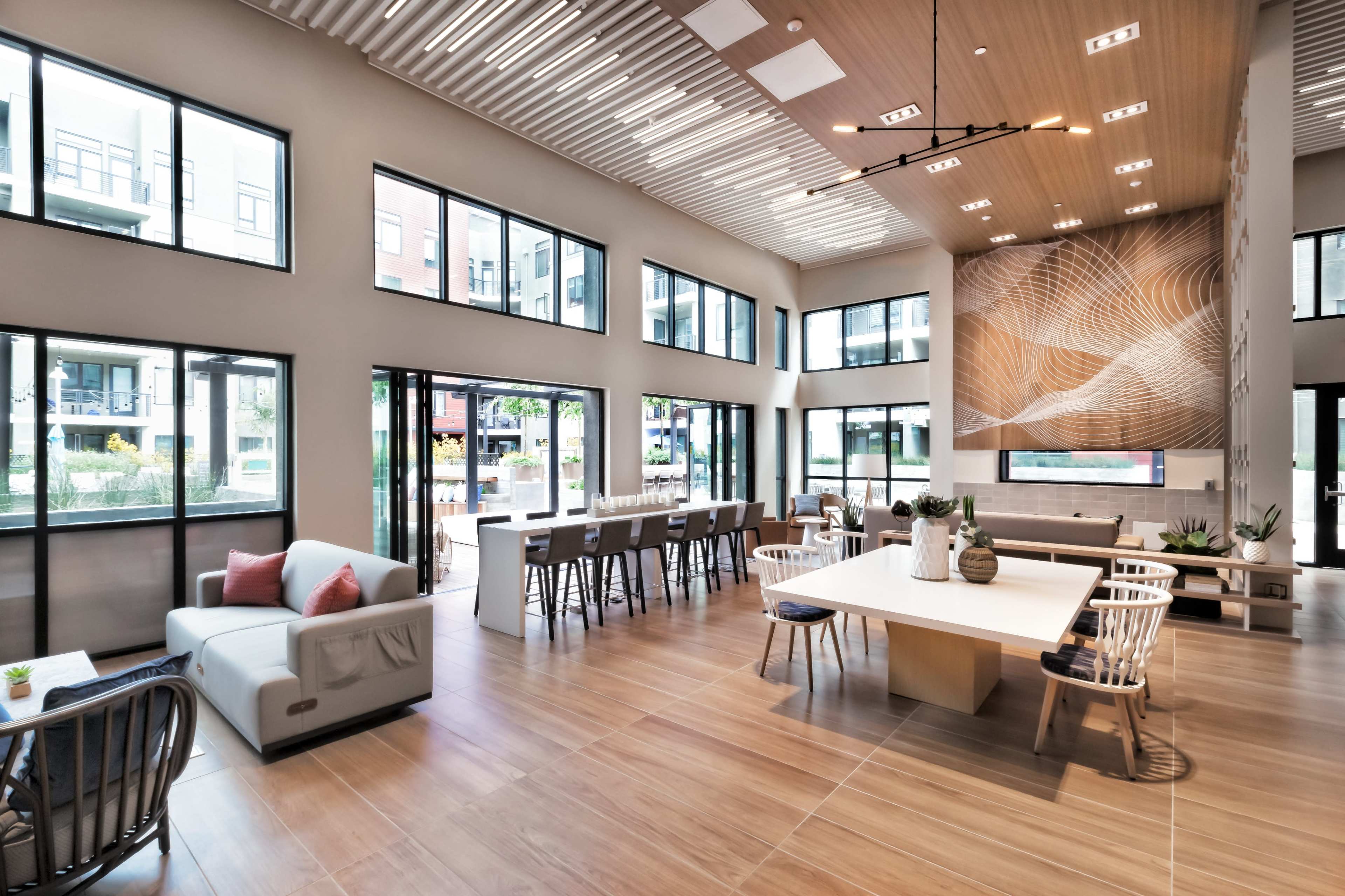 A spacious and modern communal living area with large windows, light wooden accents, and a mix of seating arrangements including a dining table, couches, and bar stools.
