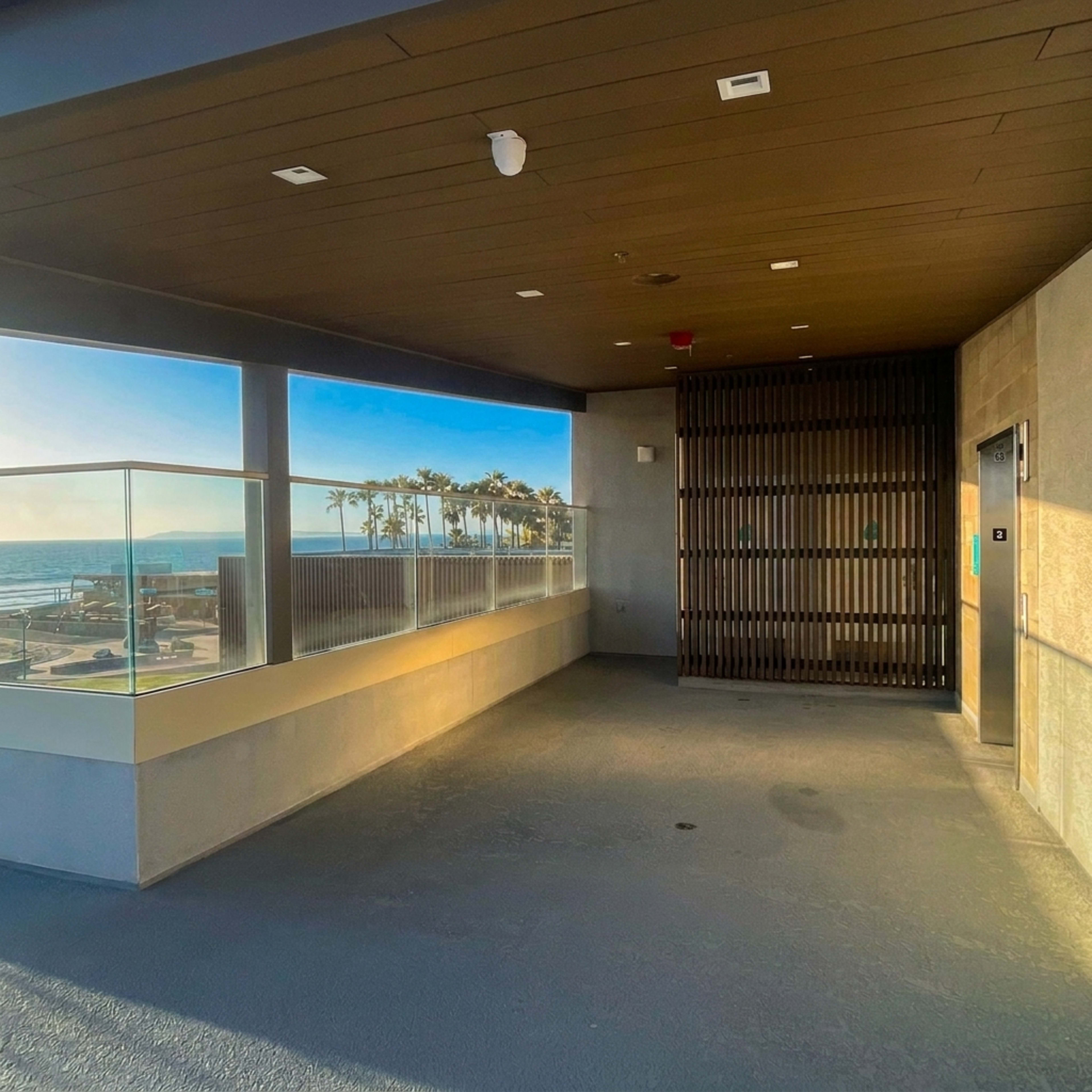 The image shows a modern, empty terrace area with a glass railing overlooking a seaside view and palm trees.