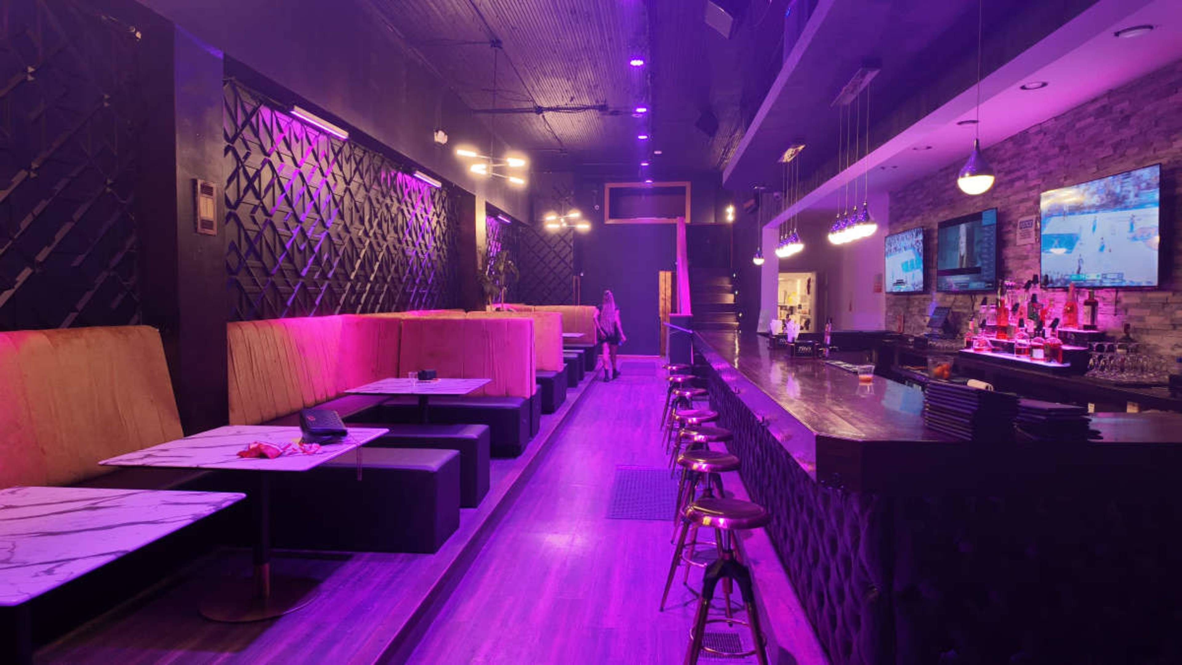 The image shows a dimly lit bar interior with plush seating and a long countertop, featuring purple lighting and multiple television screens.