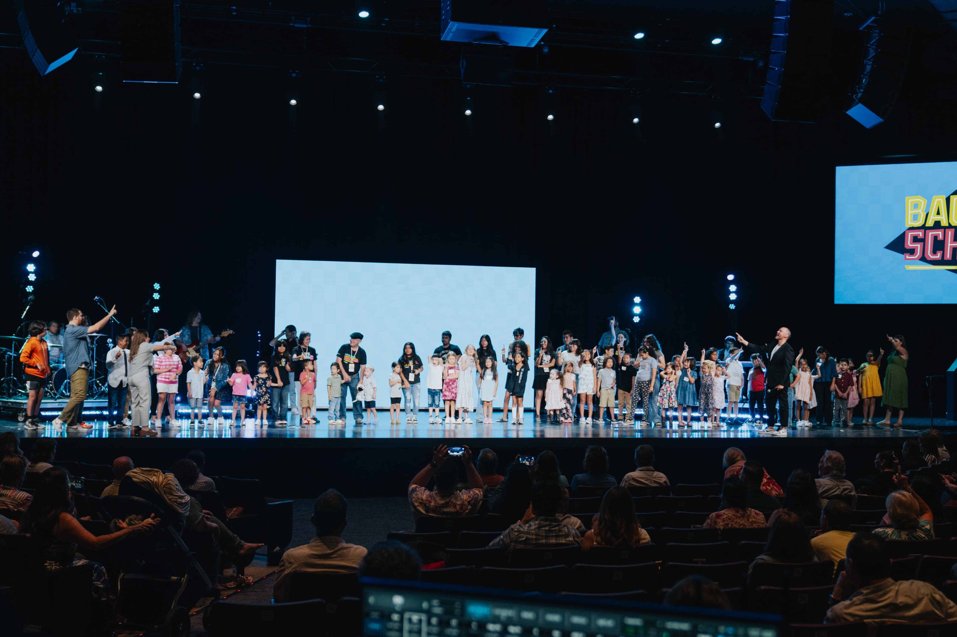 A large group of children and adults stands on stage, facing an audience, with bright stage lights and a blank screen in the background.