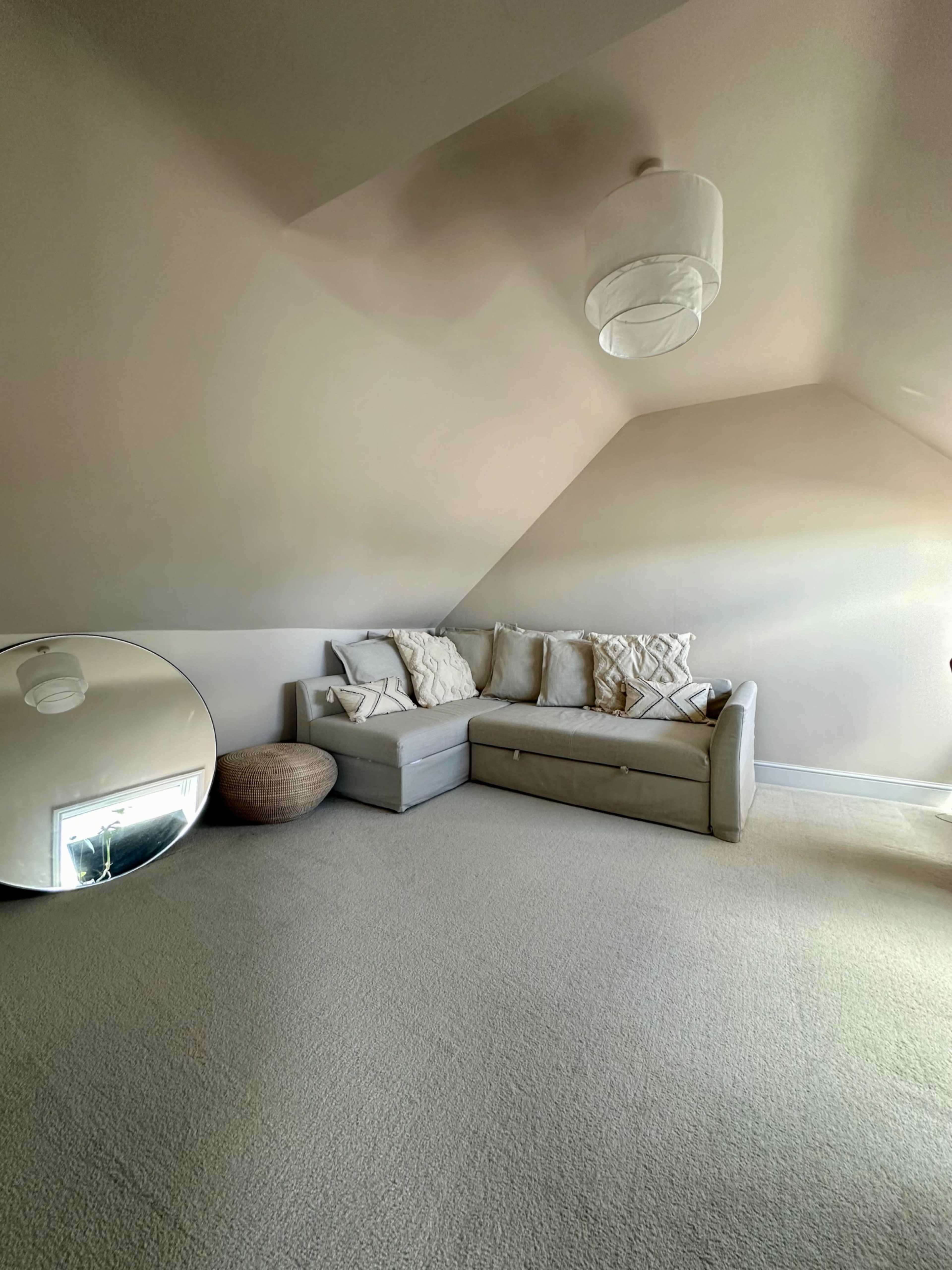 The image shows a minimalist attic room featuring a light-colored couch with cushions, a round mirror, and a woven basket.