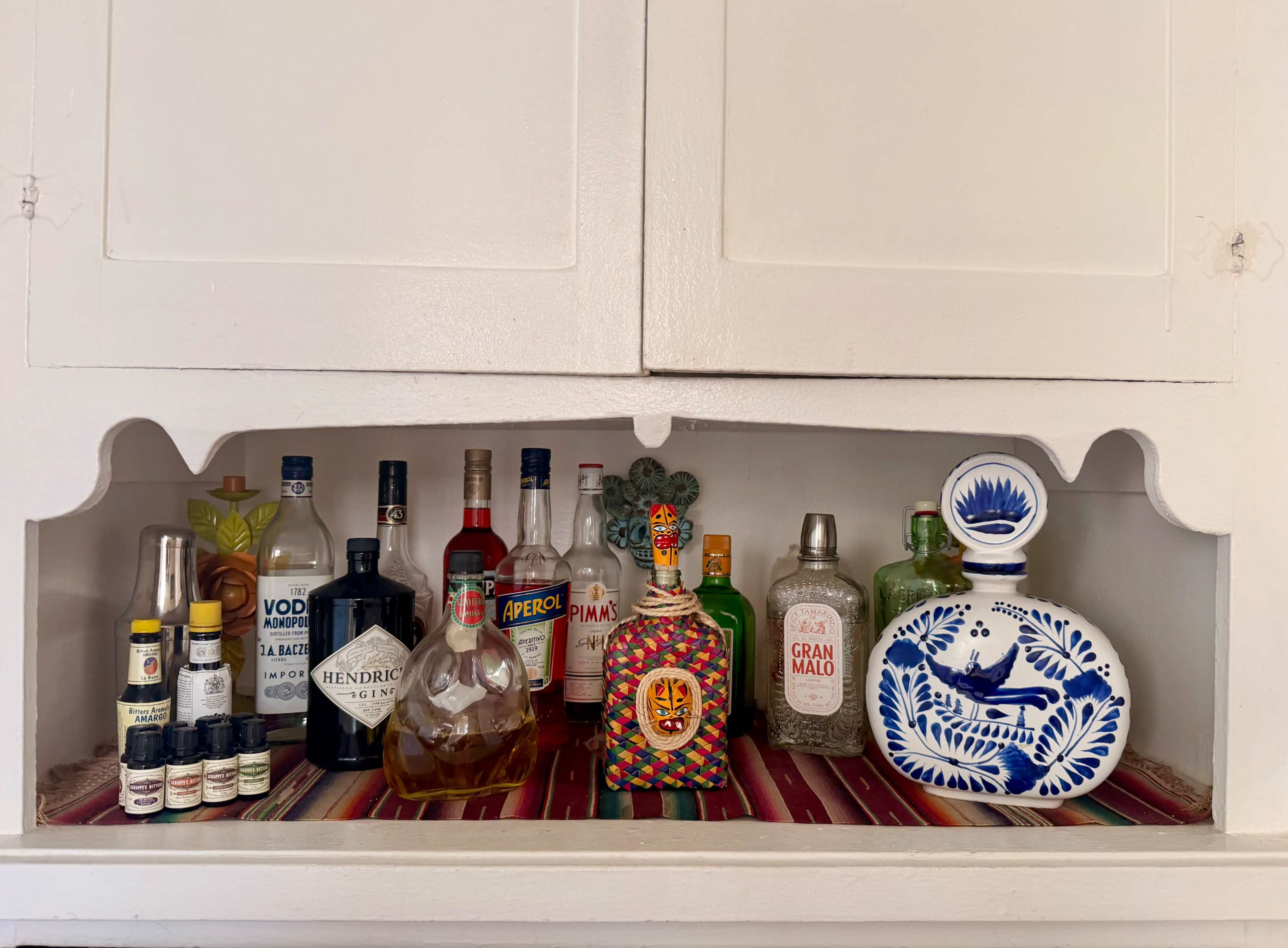 A selection of various bottles of liquor and bitters is arranged on a shelf with a decorative textile below.