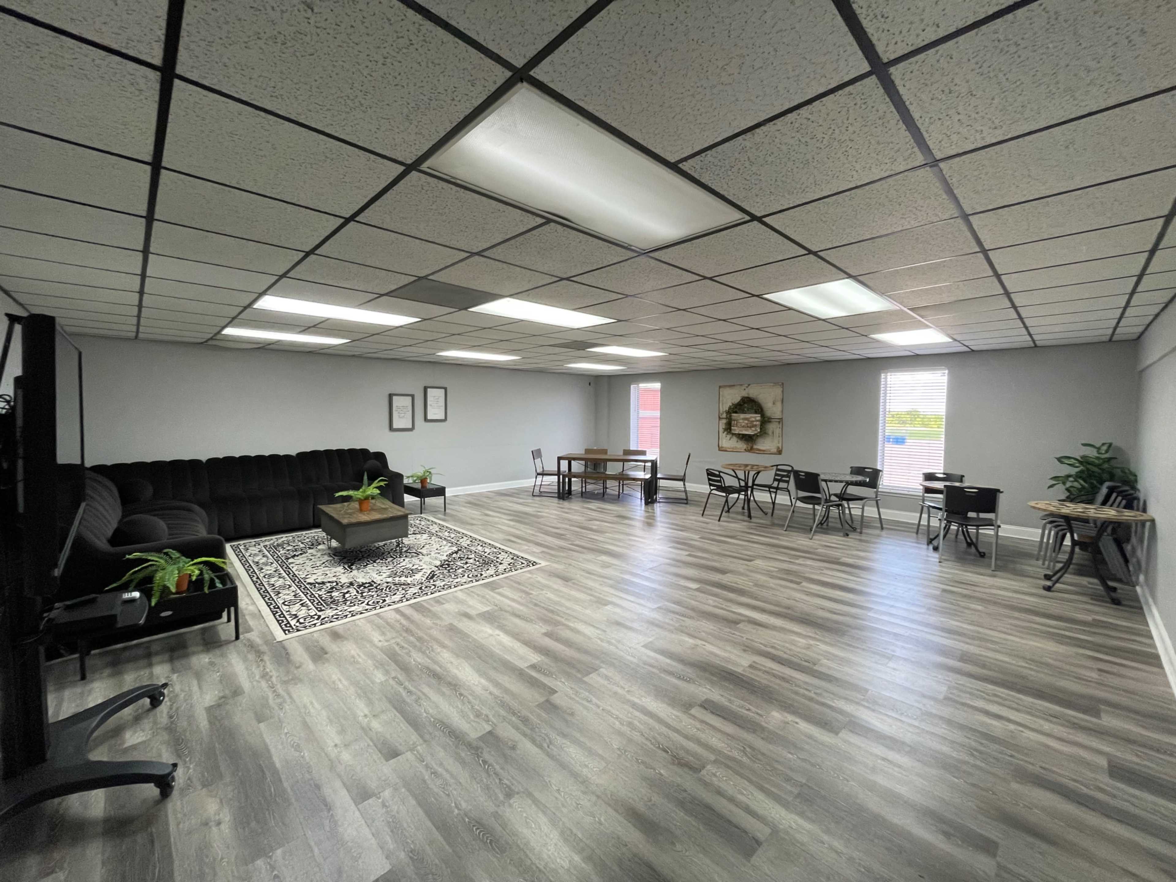 The image shows a spacious room with a dark couch, a table, several chairs, and plants, all set on a gray floor with ceiling tiles and bright overhead lights.