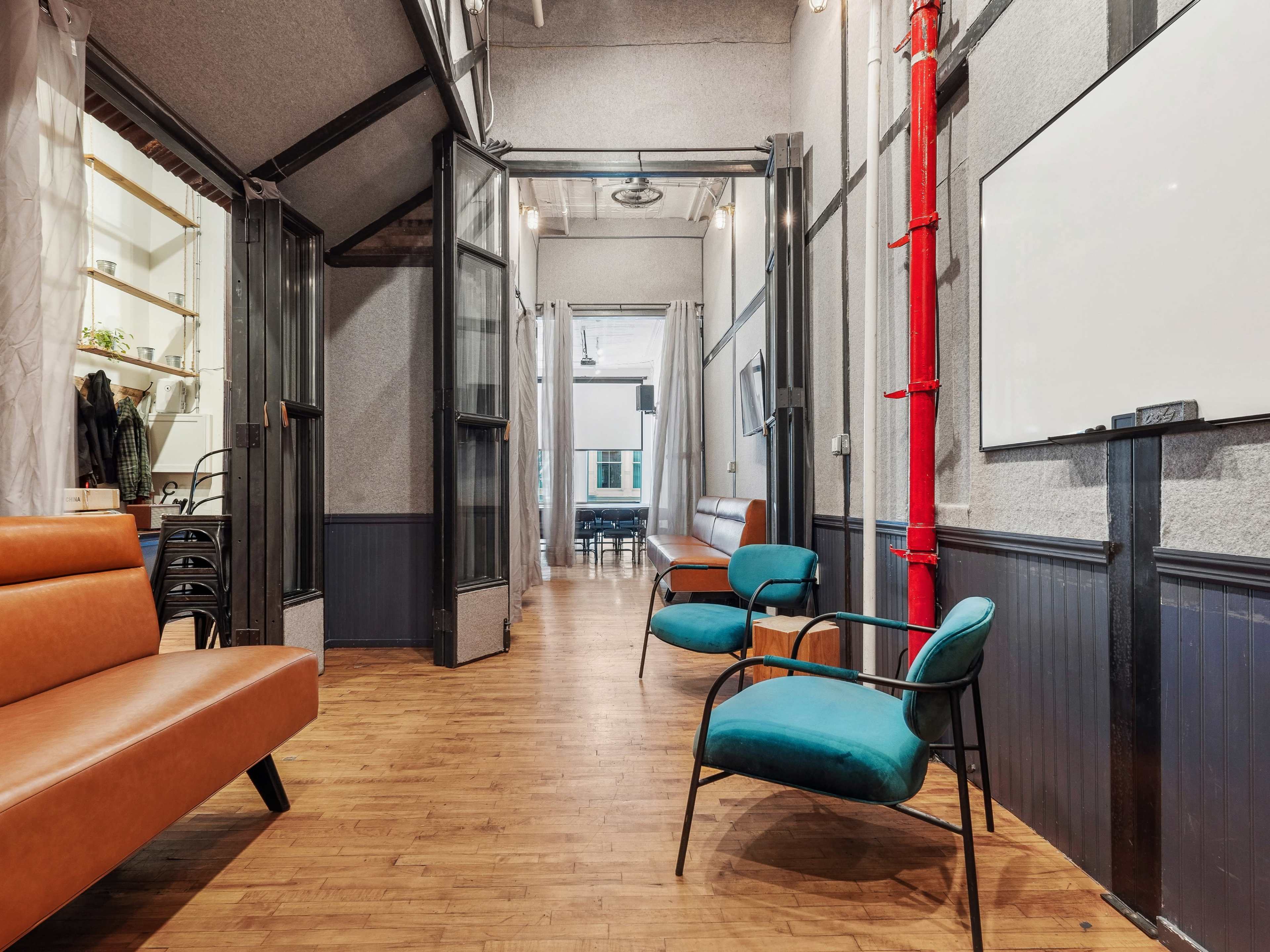 The image depicts a modern interior hallway featuring a mix of seating options, including a brown sofa and teal chairs, along with a blend of gray walls and wooden flooring.