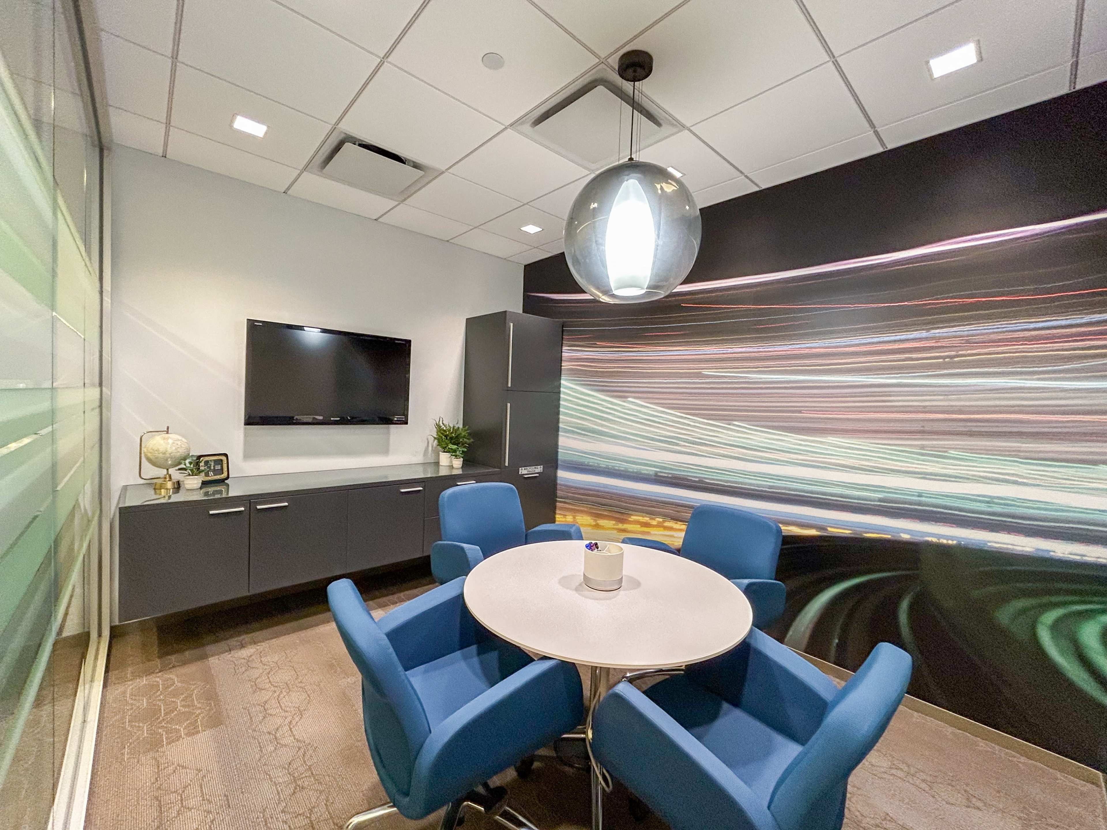 A small conference room features a circular table surrounded by four blue chairs, a wall-mounted television, and a large graphic mural of light trails.