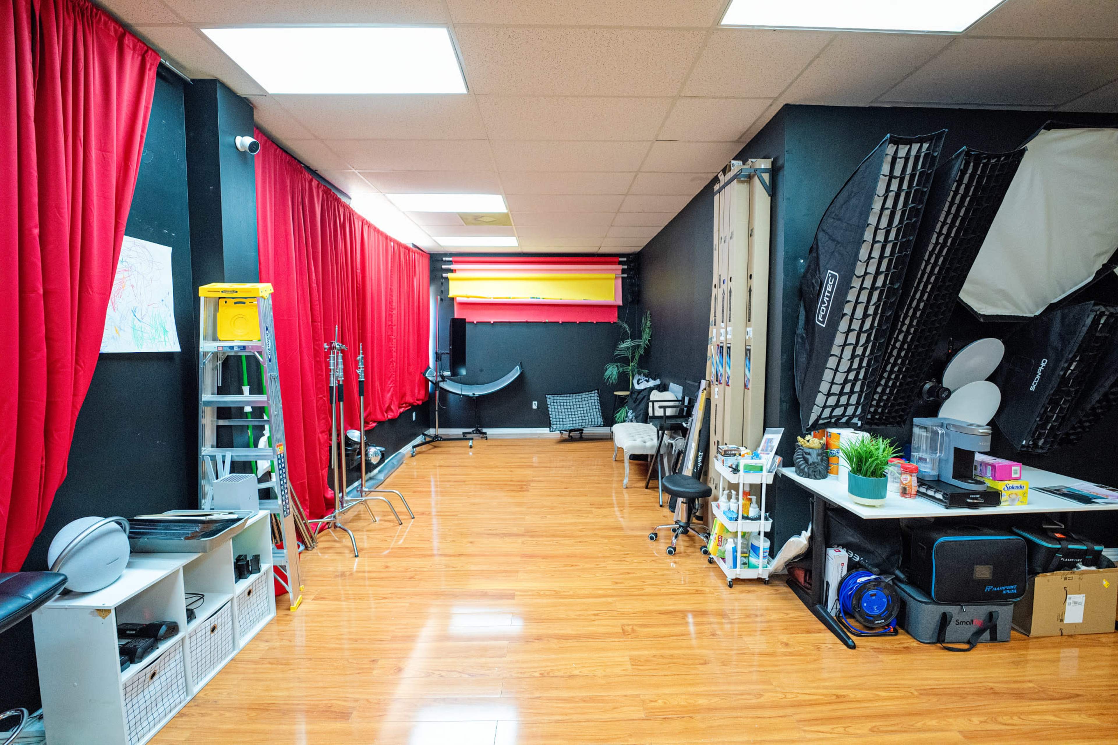 The image shows a photography studio with red curtains lining one wall, various equipment, and a polished wooden floor.