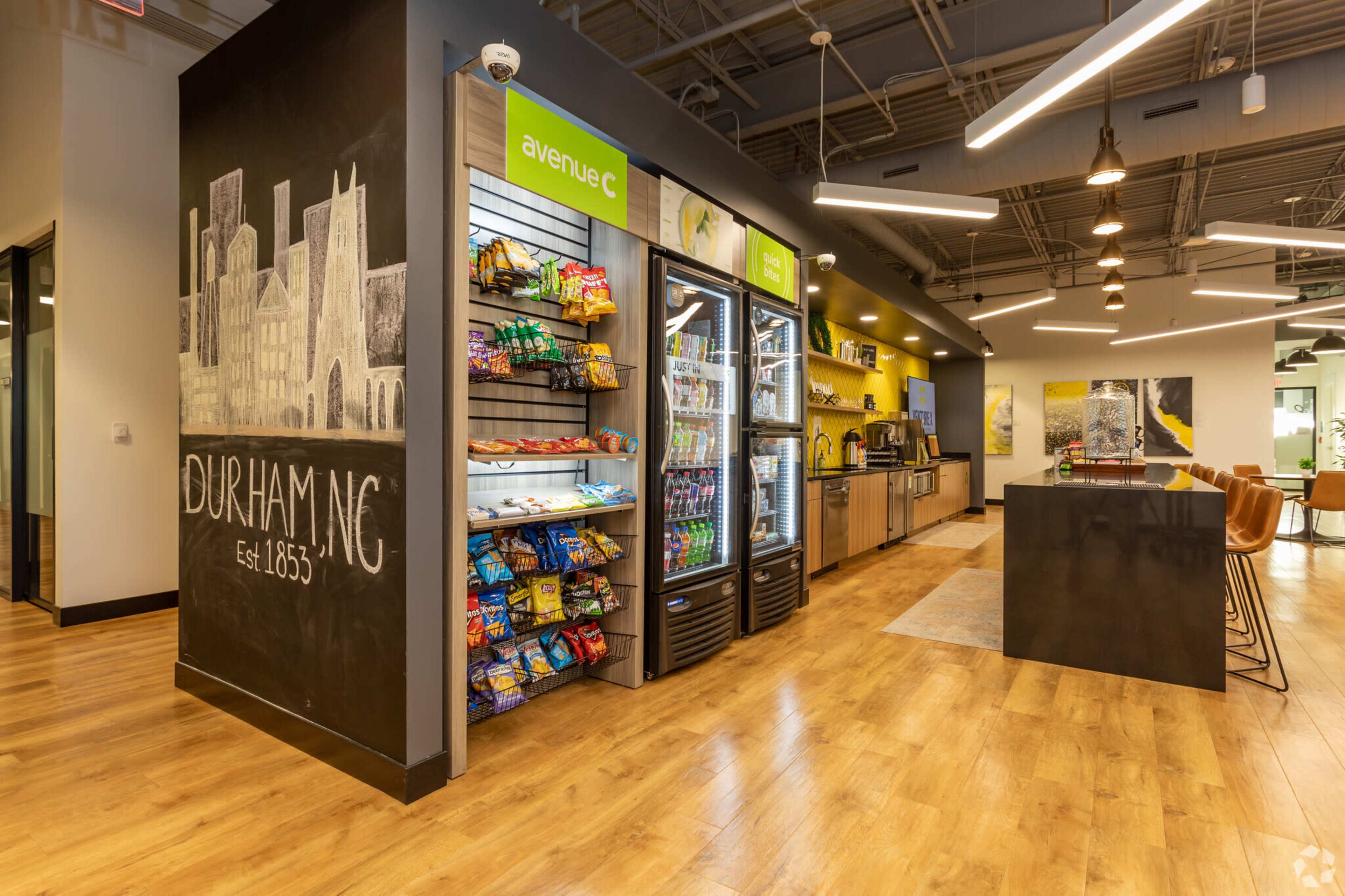 The image shows a modern office break room with a snack wall, a refrigerator, and a coffee station, featuring the text "DURHAM, NC Est. 1865" on the wall.