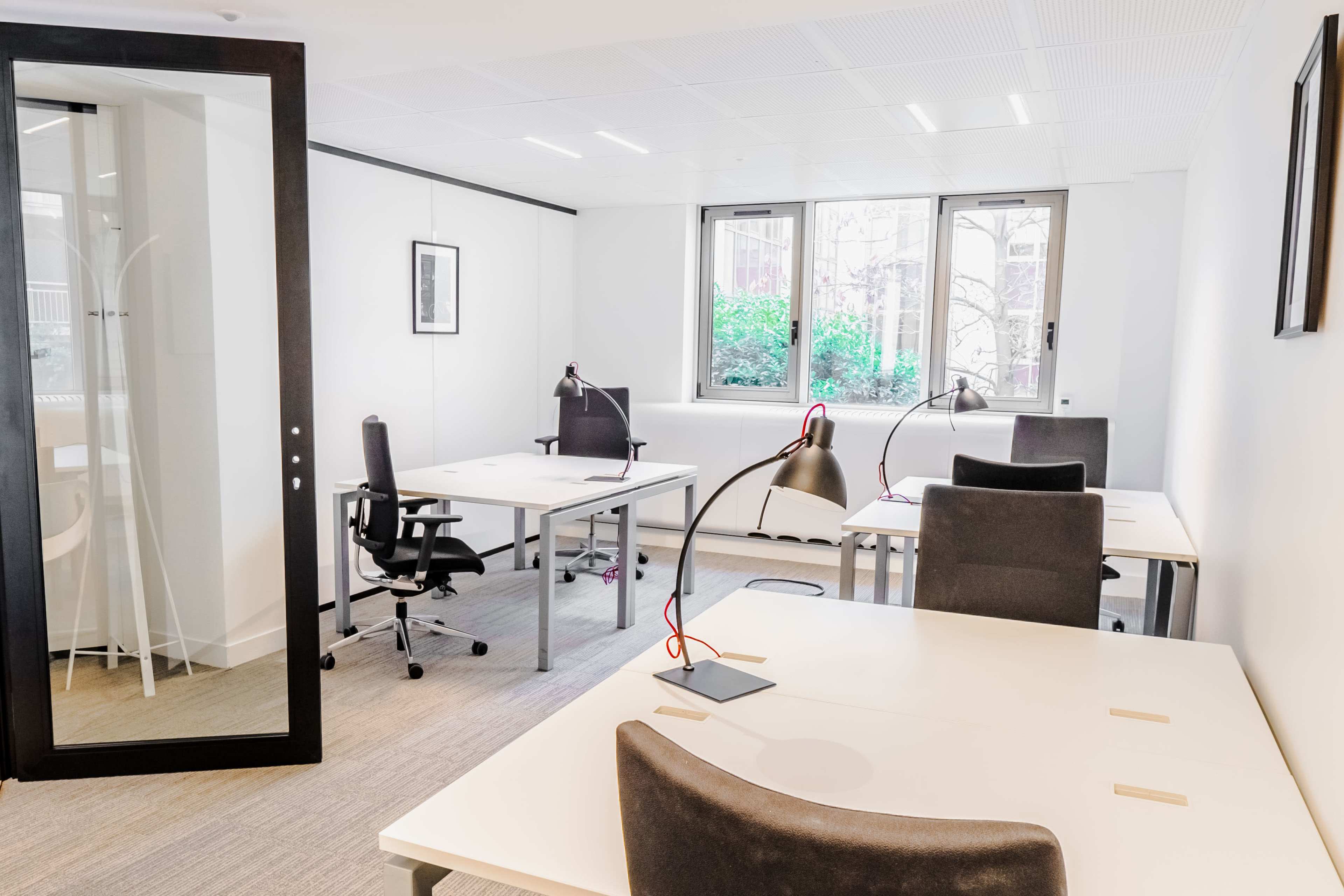 The image shows a modern office space with four desks, each equipped with a lamp, and a glass partitioned area in the background.
