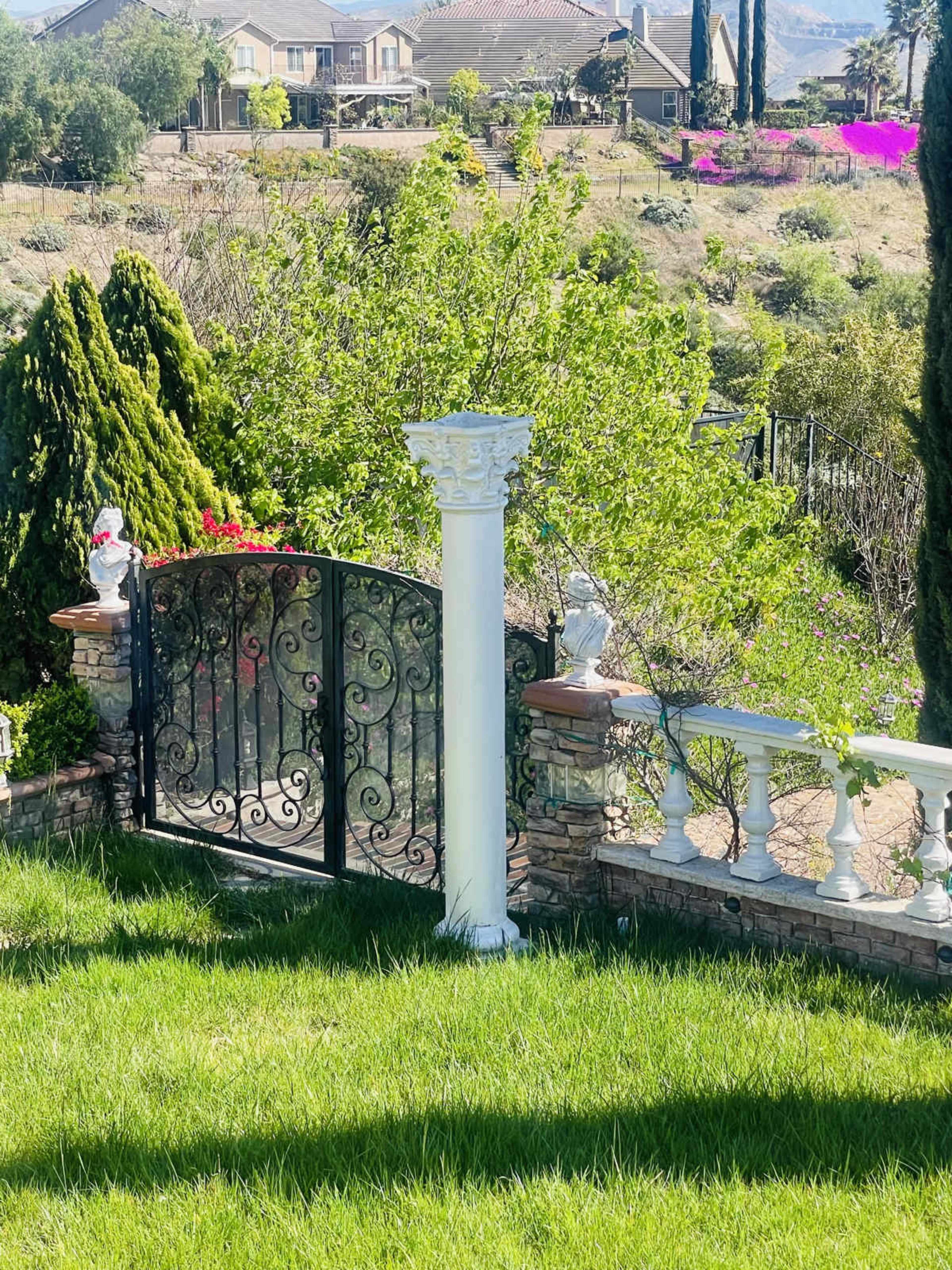 A decorative white column stands beside an ornate black metal gate, with greenery and homes visible in the background.