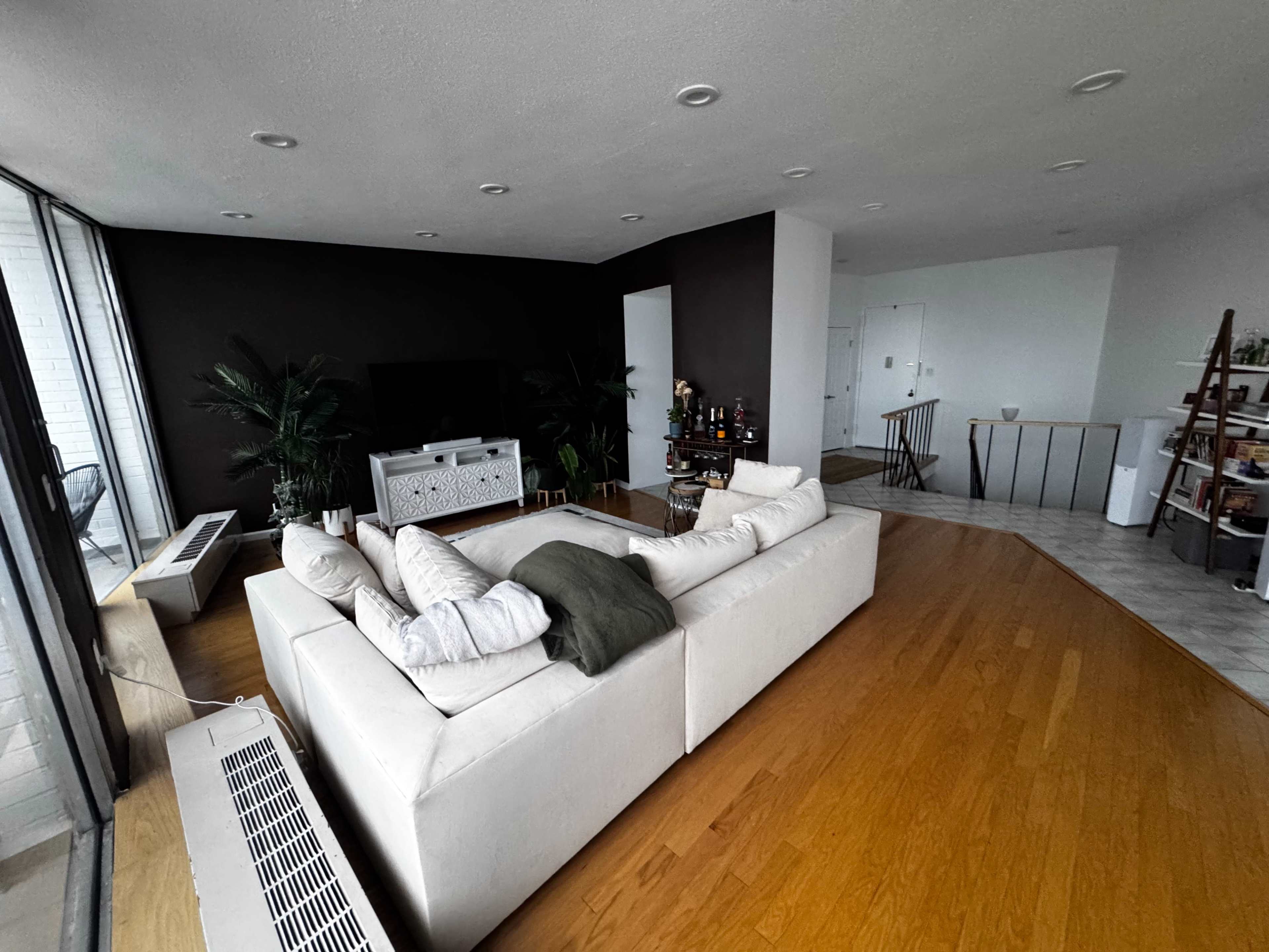 The image shows a modern living room with a large sectional sofa, a television unit, and a bar area, featuring hardwood flooring and large windows letting in natural light.