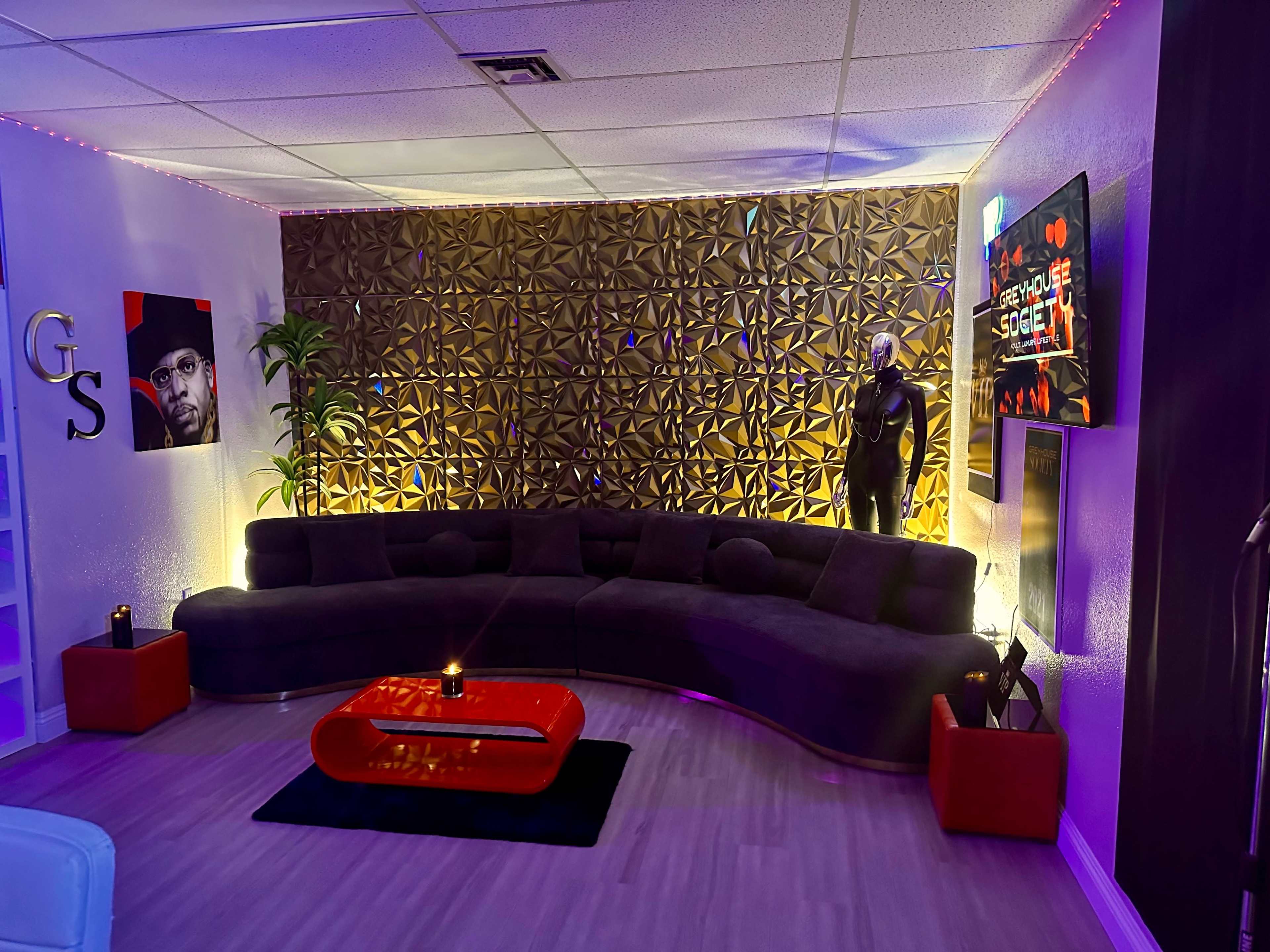 A modern lounge area with a curved gray sofa, a red coffee table, a decorative wall with geometric patterns, and a TV mounted on the wall.