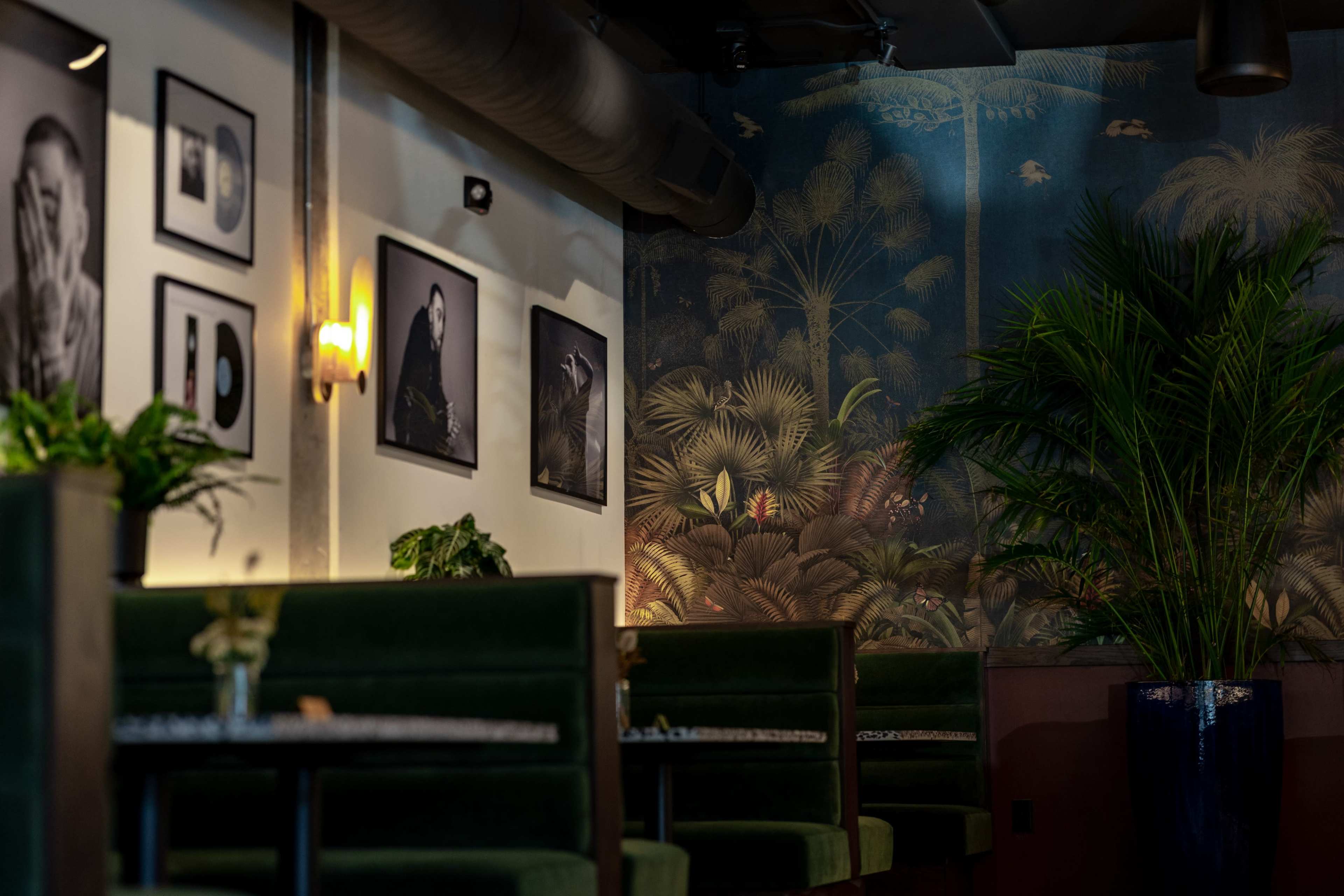 The image shows a stylish restaurant interior featuring green booths, framed black-and-white photographs on the walls, and a large mural of tropical plants.