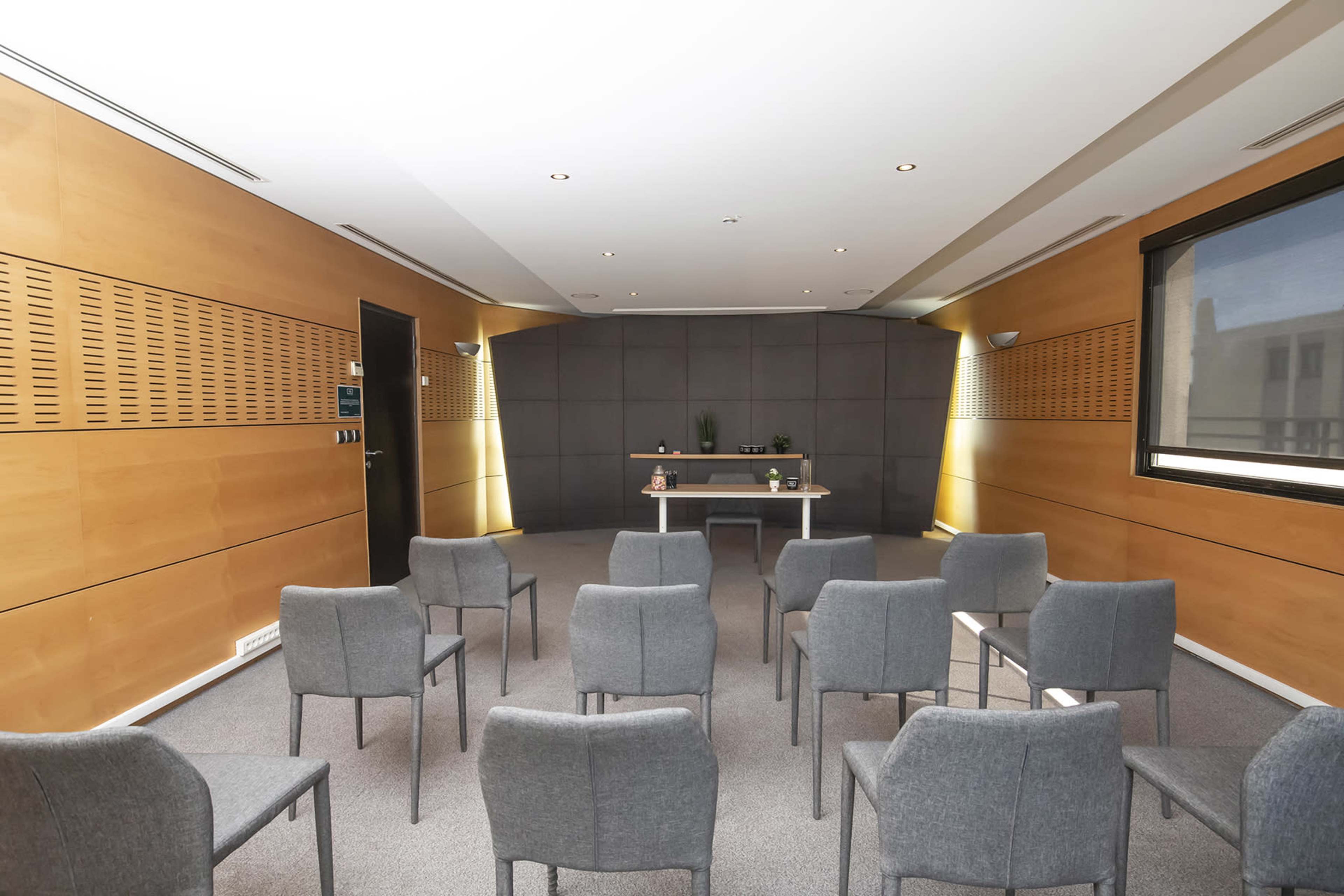 A conference room is arranged with a row of gray chairs facing a central table against a neutral backdrop.