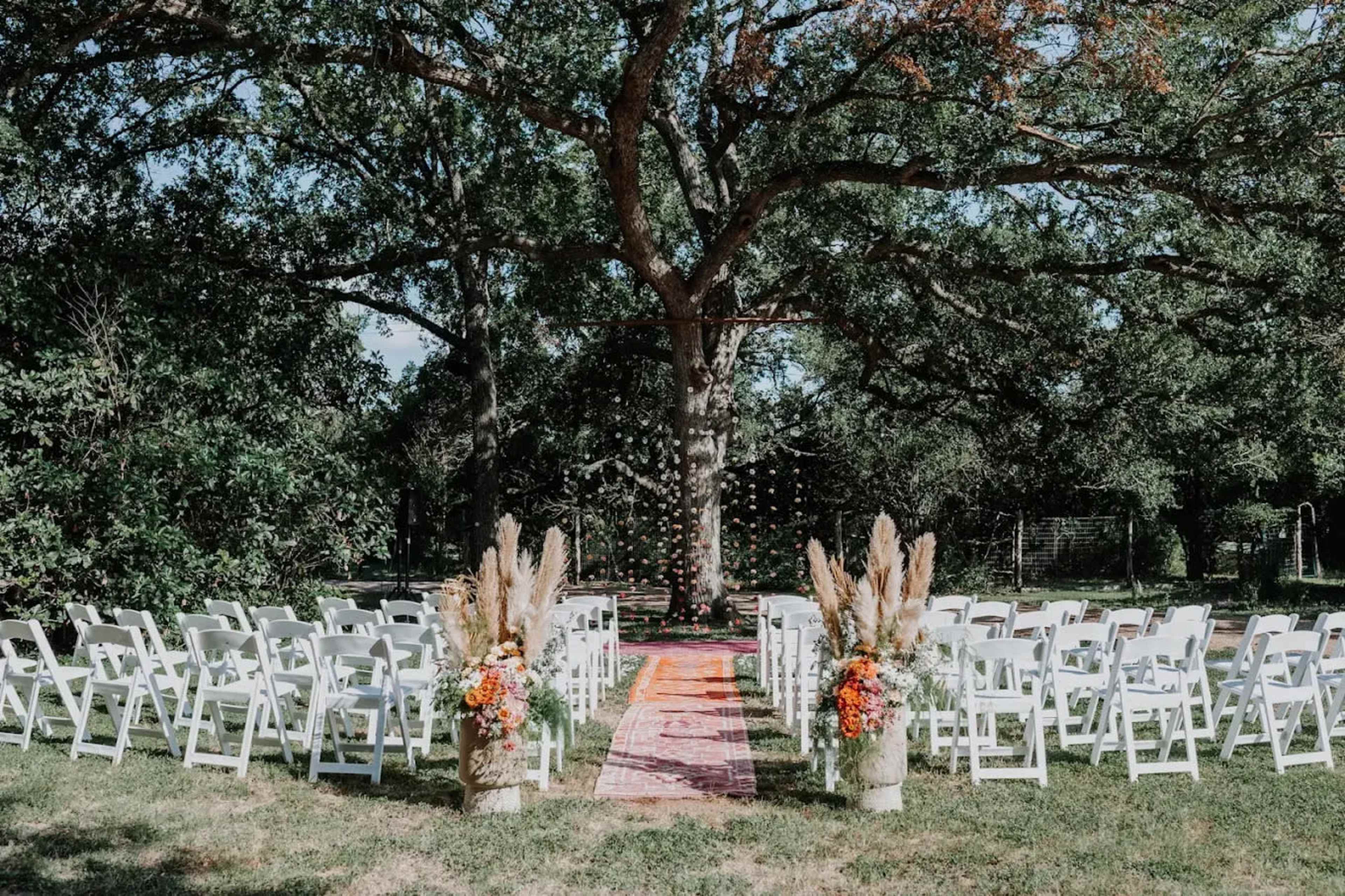 A wedding ceremony setup features white chairs arranged in rows on a grassy area under a large tree, with a colorful rug leading to a decorative backdrop adorned with floral accents.