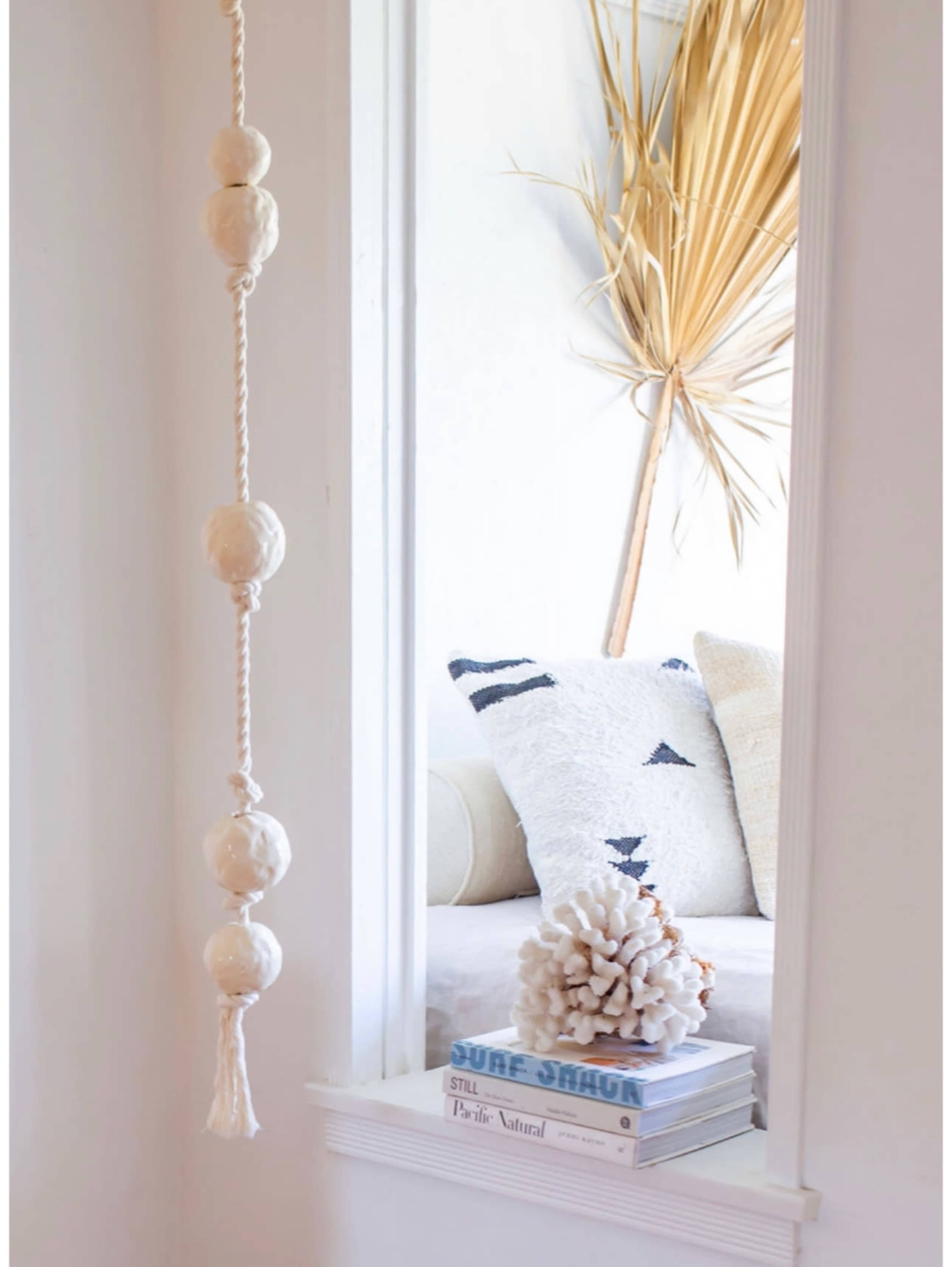 The image showcases a bright interior featuring a mirror with a decorative rope and bead hanging, flanked by decorative pillows and a large dried palm leaf.