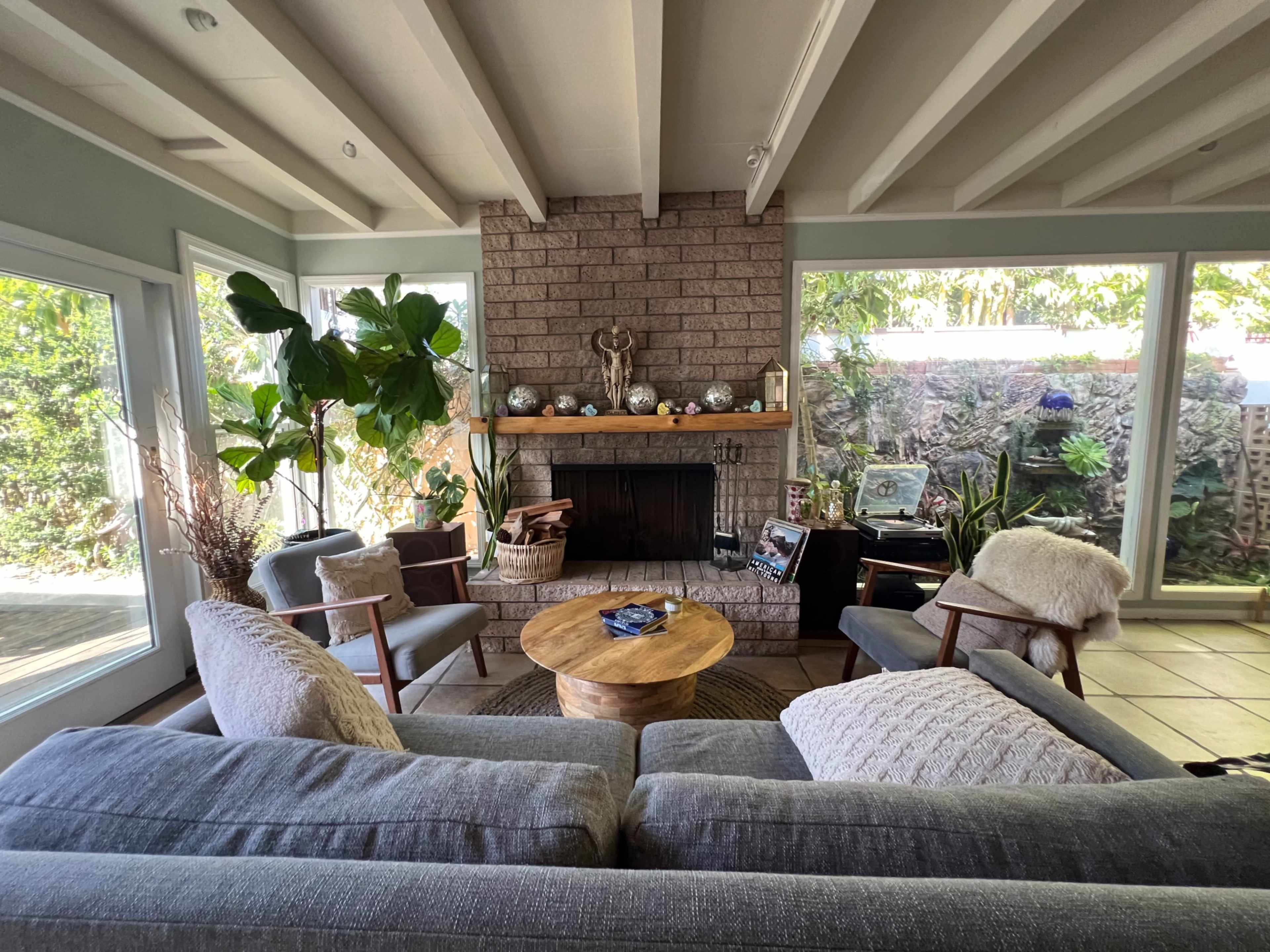 A cozy living room features a stone fireplace, a circular wooden coffee table, and comfortable seating arranged around large windows with views of greenery.