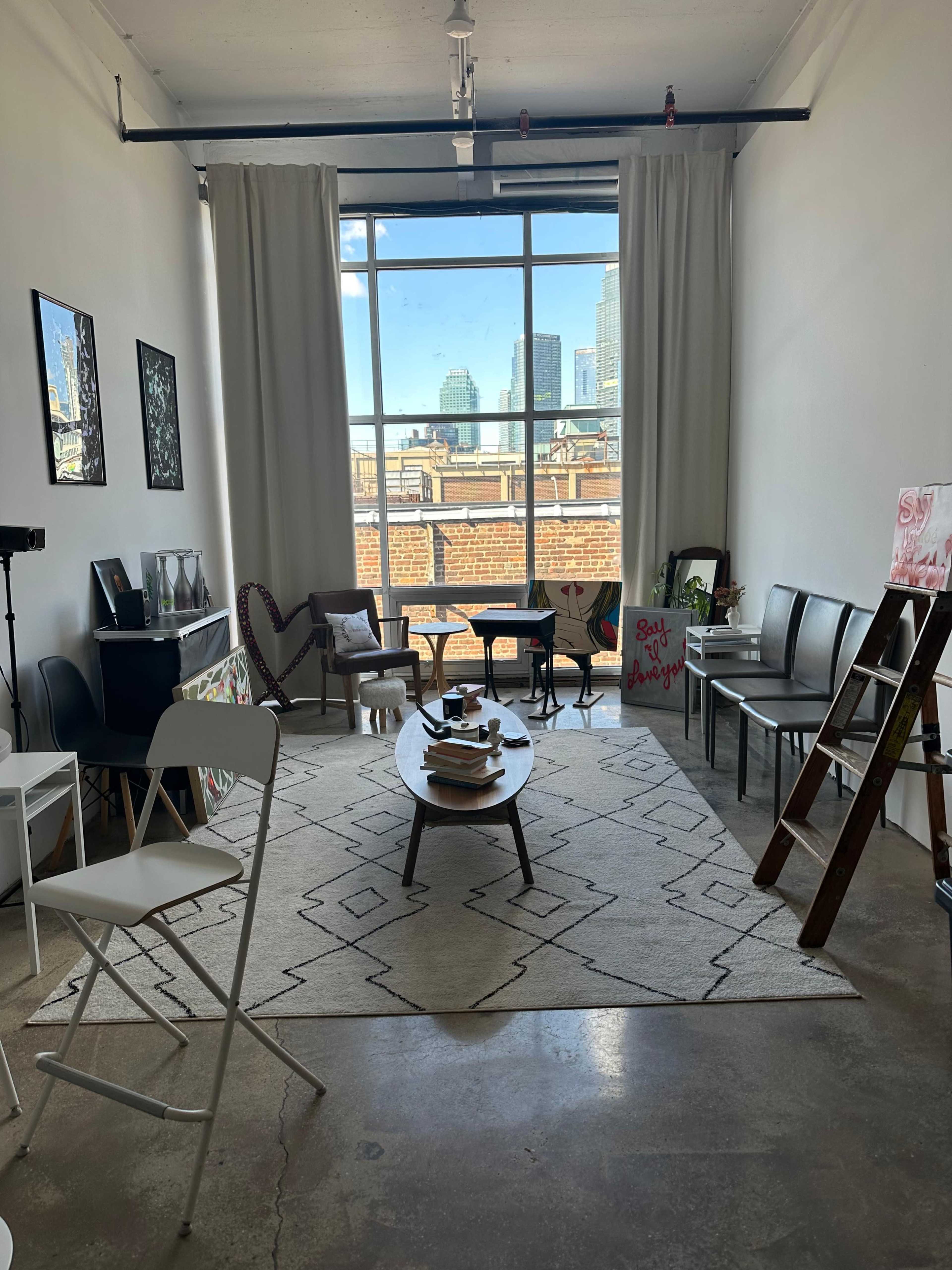 The image shows a bright, modern room with large windows, a patterned rug on the floor, chairs arranged around a central table, and various pieces of artwork on the walls.